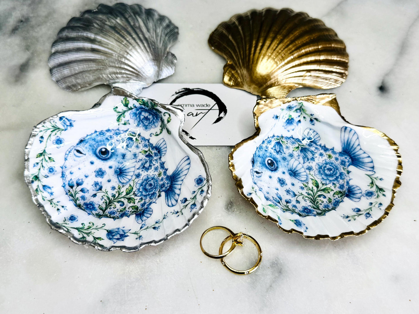 Handcrafted scallop shell trinket dish featuring an original Delft Blue puffer fish illustration created and designed by Emma Wade ART. Natural scallop shell finished with a choice of genuine gold leaf or silver leaf and sealed beneath crystal-hard glossy resin. Coastal ring holder, jewellery dish and decorative shell tray inspired by traditional Delft ceramics and marine life. Available in standard 10–11cm or large 12–14cm sizes.