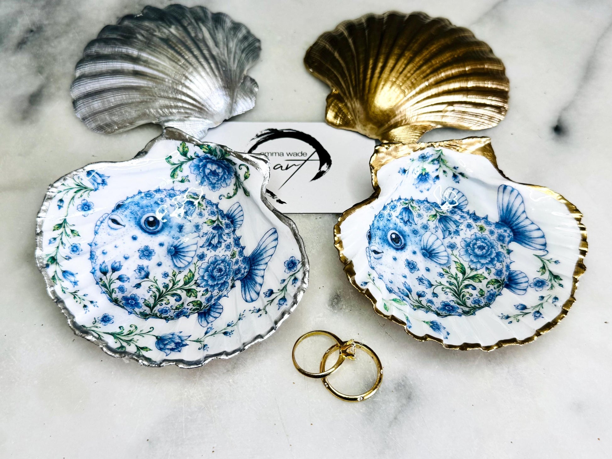 Handcrafted scallop shell trinket dish featuring an original Delft Blue puffer fish illustration created and designed by Emma Wade ART. Natural scallop shell finished with a choice of genuine gold leaf or silver leaf and sealed beneath crystal-hard glossy resin. Coastal ring holder, jewellery dish and decorative shell tray inspired by traditional Delft ceramics and marine life. Available in standard 10–11cm or large 12–14cm sizes.