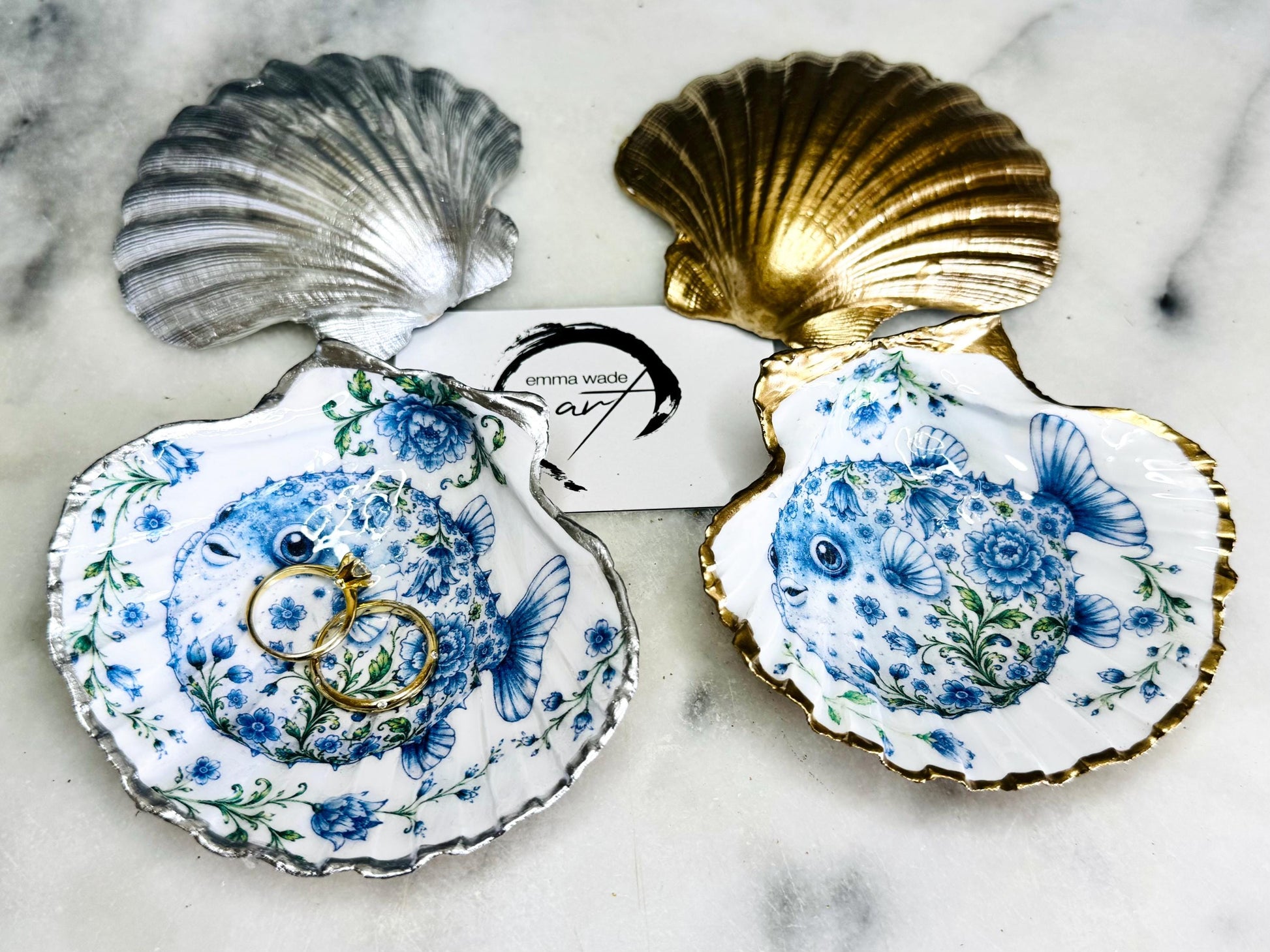 Handcrafted scallop shell trinket dish featuring an original Delft Blue puffer fish illustration created and designed by Emma Wade ART. Natural scallop shell finished with a choice of genuine gold leaf or silver leaf and sealed beneath crystal-hard glossy resin. Coastal ring holder, jewellery dish and decorative shell tray inspired by traditional Delft ceramics and marine life. Available in standard 10–11cm or large 12–14cm sizes.