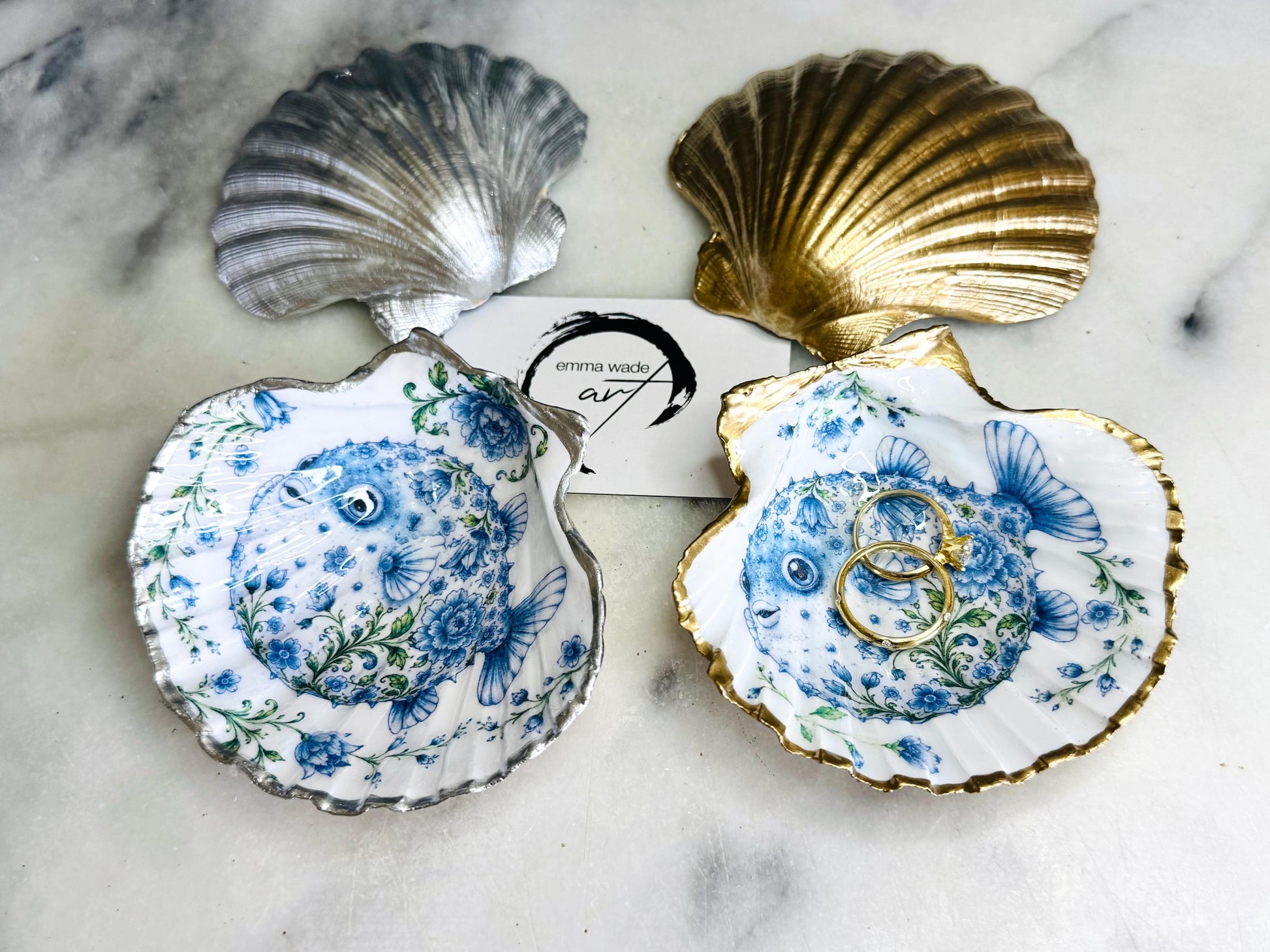 Handcrafted scallop shell trinket dish featuring an original Delft Blue puffer fish illustration created and designed by Emma Wade ART. Natural scallop shell finished with a choice of genuine gold leaf or silver leaf and sealed beneath crystal-hard glossy resin. Coastal ring holder, jewellery dish and decorative shell tray inspired by traditional Delft ceramics and marine life. Available in standard 10–11cm or large 12–14cm sizes.