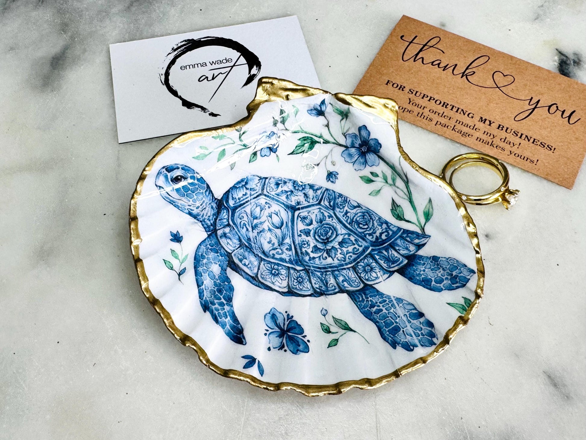 Handcrafted scallop shell trinket dish featuring an original illustrated Delft Blue Sea Turtle design by Emma Wade ART. Natural shell with hand-gilded gold or silver leaf rim and glossy resin finish. Coastal jewellery tray for rings, earrings and keepsakes. Beach house decor, sea life gift and ocean-inspired home accessory available in standard and large sizes.