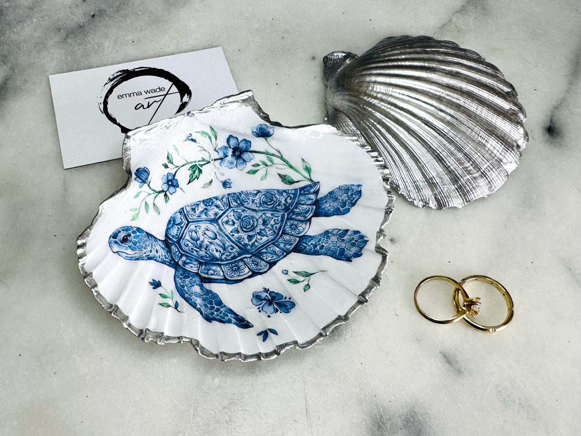 Handcrafted scallop shell trinket dish featuring an original illustrated Delft Blue Sea Turtle design by Emma Wade ART. Natural shell with hand-gilded gold or silver leaf rim and glossy resin finish. Coastal jewellery tray for rings, earrings and keepsakes. Beach house decor, sea life gift and ocean-inspired home accessory available in standard and large sizes.