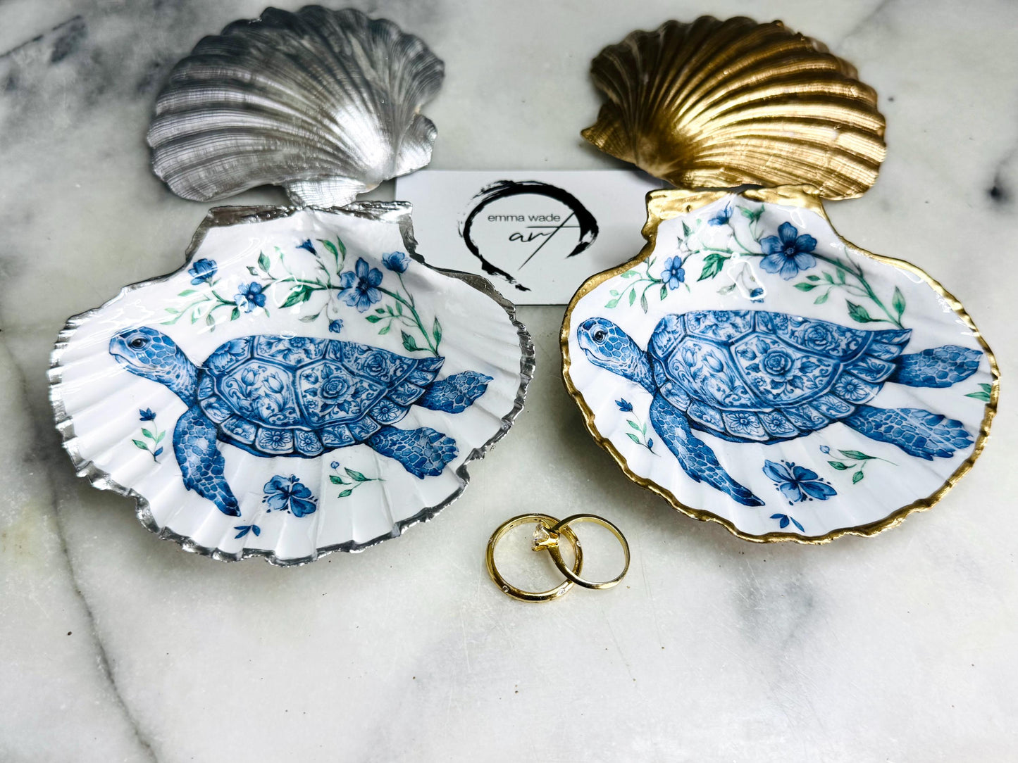 Handcrafted scallop shell trinket dish featuring an original illustrated Delft Blue Sea Turtle design by Emma Wade ART. Natural shell with hand-gilded gold or silver leaf rim and glossy resin finish. Coastal jewellery tray for rings, earrings and keepsakes. Beach house decor, sea life gift and ocean-inspired home accessory available in standard and large sizes.
