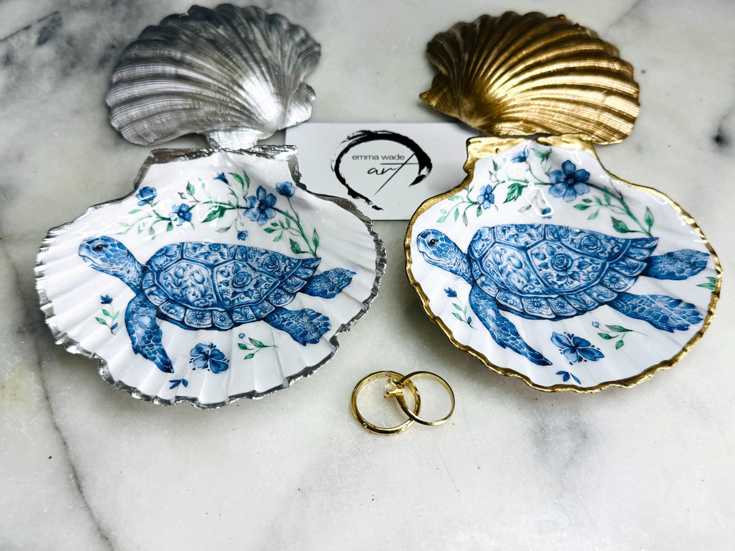 Handcrafted scallop shell trinket dish featuring an original illustrated Delft Blue Sea Turtle design by Emma Wade ART. Natural shell with hand-gilded gold or silver leaf rim and glossy resin finish. Coastal jewellery tray for rings, earrings and keepsakes. Beach house decor, sea life gift and ocean-inspired home accessory available in standard and large sizes.