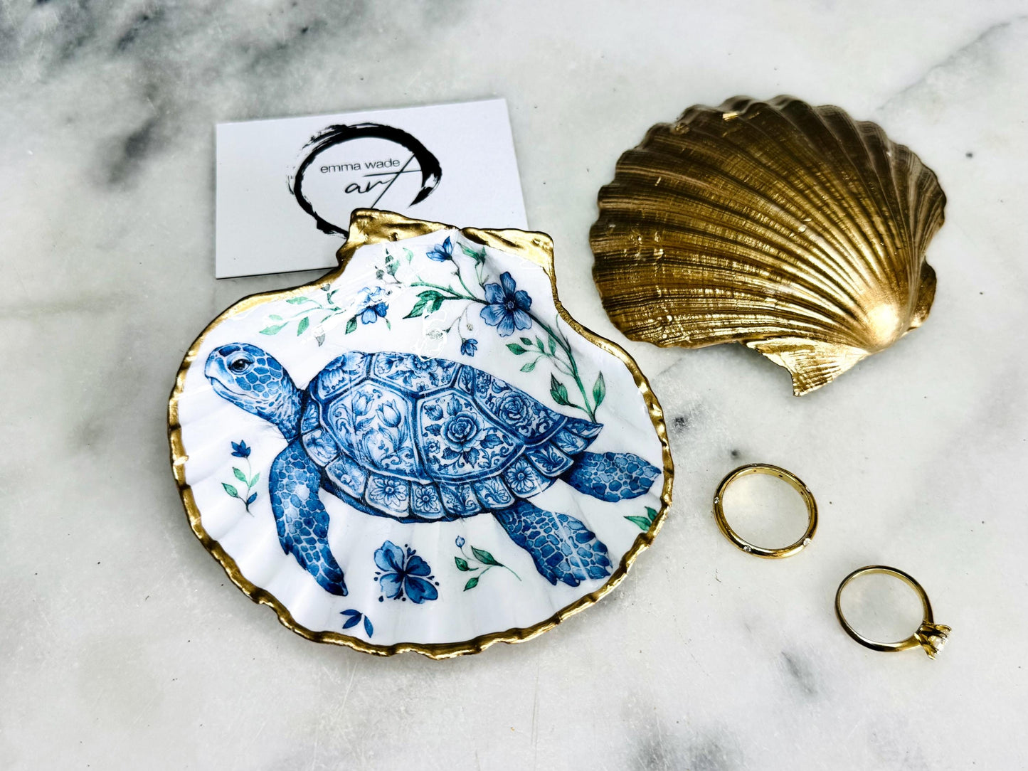 Handcrafted scallop shell trinket dish featuring an original illustrated Delft Blue Sea Turtle design by Emma Wade ART. Natural shell with hand-gilded gold or silver leaf rim and glossy resin finish. Coastal jewellery tray for rings, earrings and keepsakes. Beach house decor, sea life gift and ocean-inspired home accessory available in standard and large sizes.