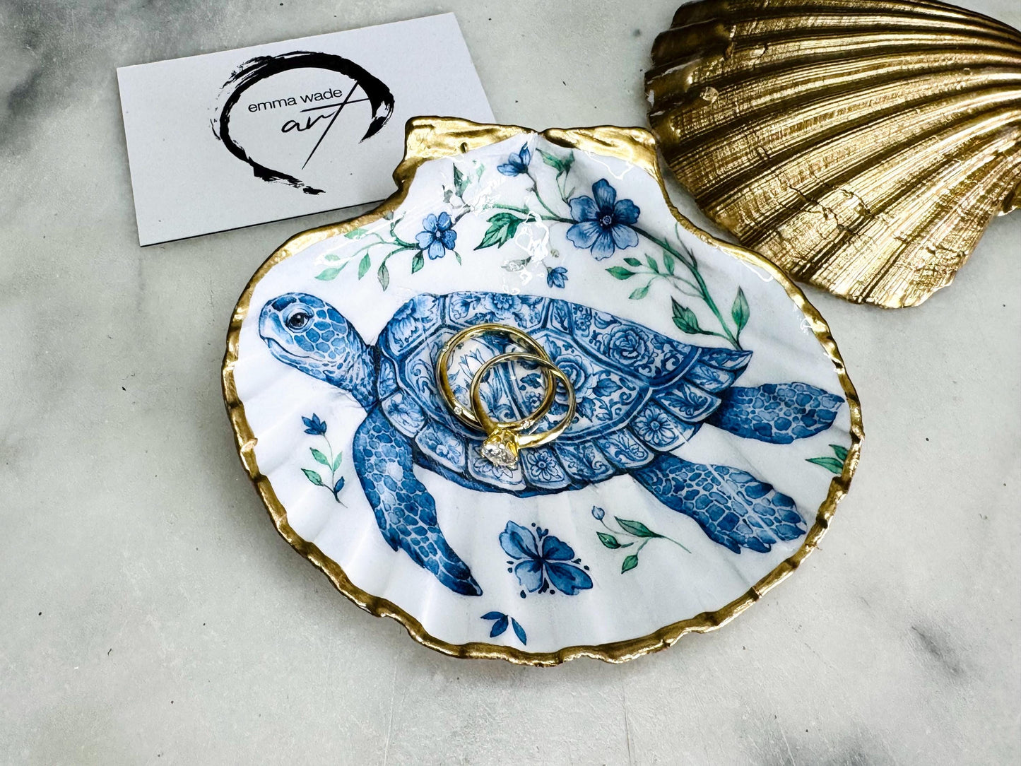 Handcrafted scallop shell trinket dish featuring an original illustrated Delft Blue Sea Turtle design by Emma Wade ART. Natural shell with hand-gilded gold or silver leaf rim and glossy resin finish. Coastal jewellery tray for rings, earrings and keepsakes. Beach house decor, sea life gift and ocean-inspired home accessory available in standard and large sizes.