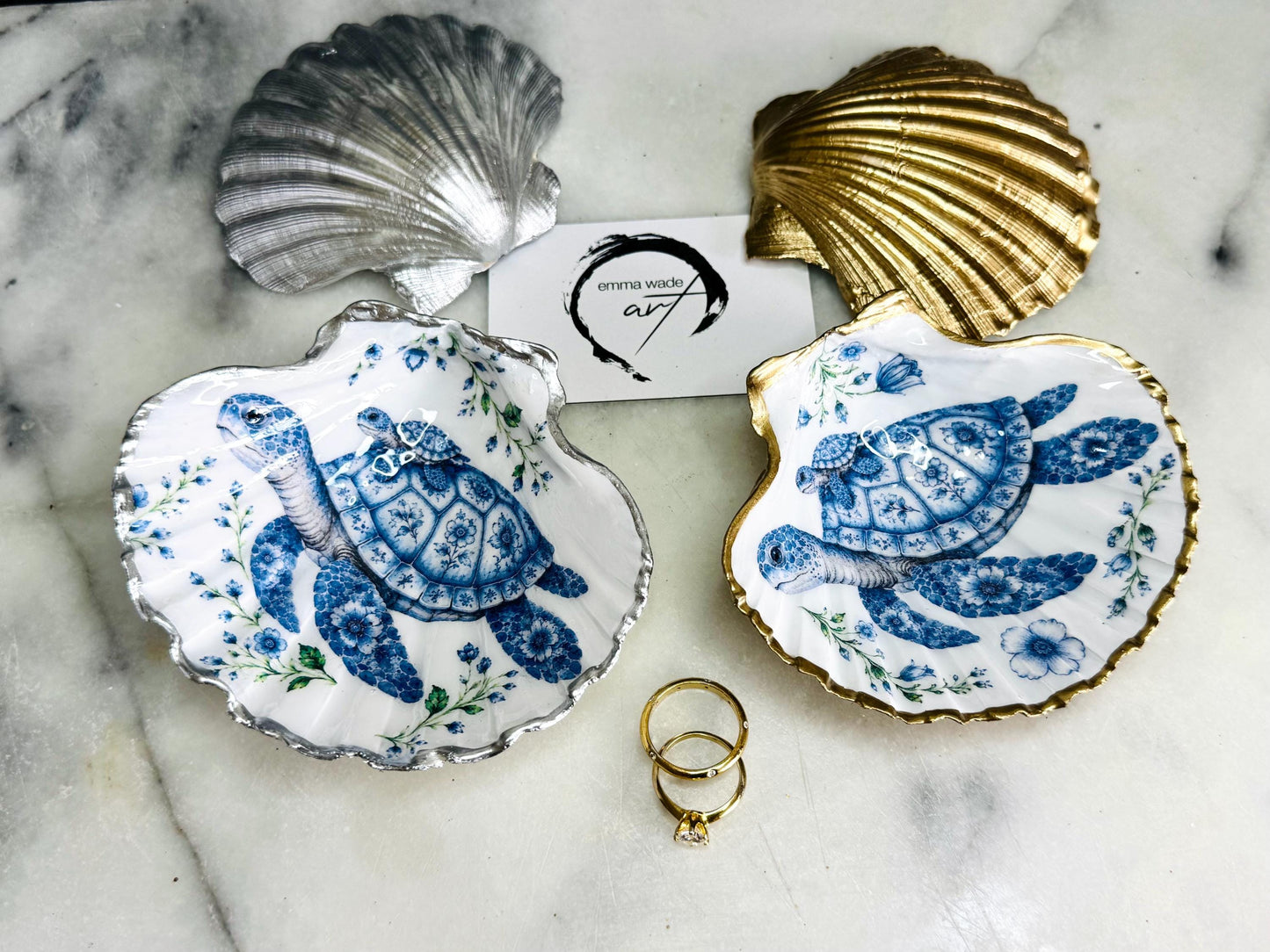 Handcrafted scallop shell trinket dish featuring an original illustrated Delft Blue Sea Turtle design by Emma Wade ART. Natural shell with hand-gilded gold or silver leaf rim and glossy resin finish. Coastal jewellery tray for rings, earrings and keepsakes. Beach house decor, sea life gift and ocean-inspired home accessory available in standard and large sizes.