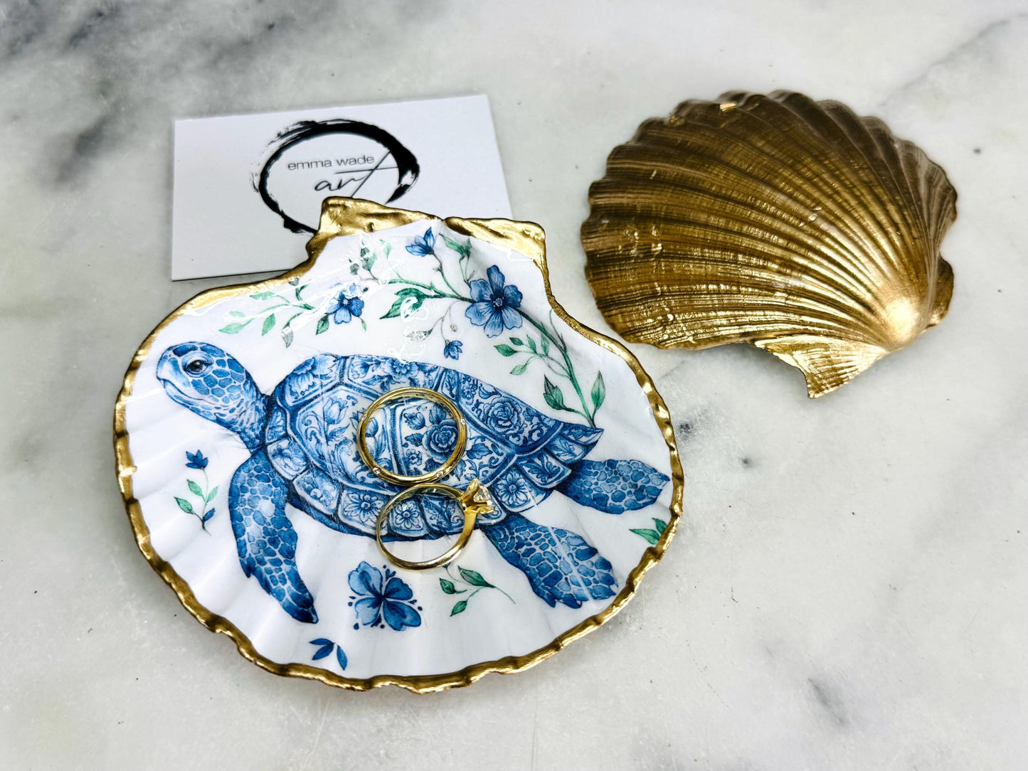 Handcrafted scallop shell trinket dish featuring an original illustrated Delft Blue Sea Turtle design by Emma Wade ART. Natural shell with hand-gilded gold or silver leaf rim and glossy resin finish. Coastal jewellery tray for rings, earrings and keepsakes. Beach house decor, sea life gift and ocean-inspired home accessory available in standard and large sizes.