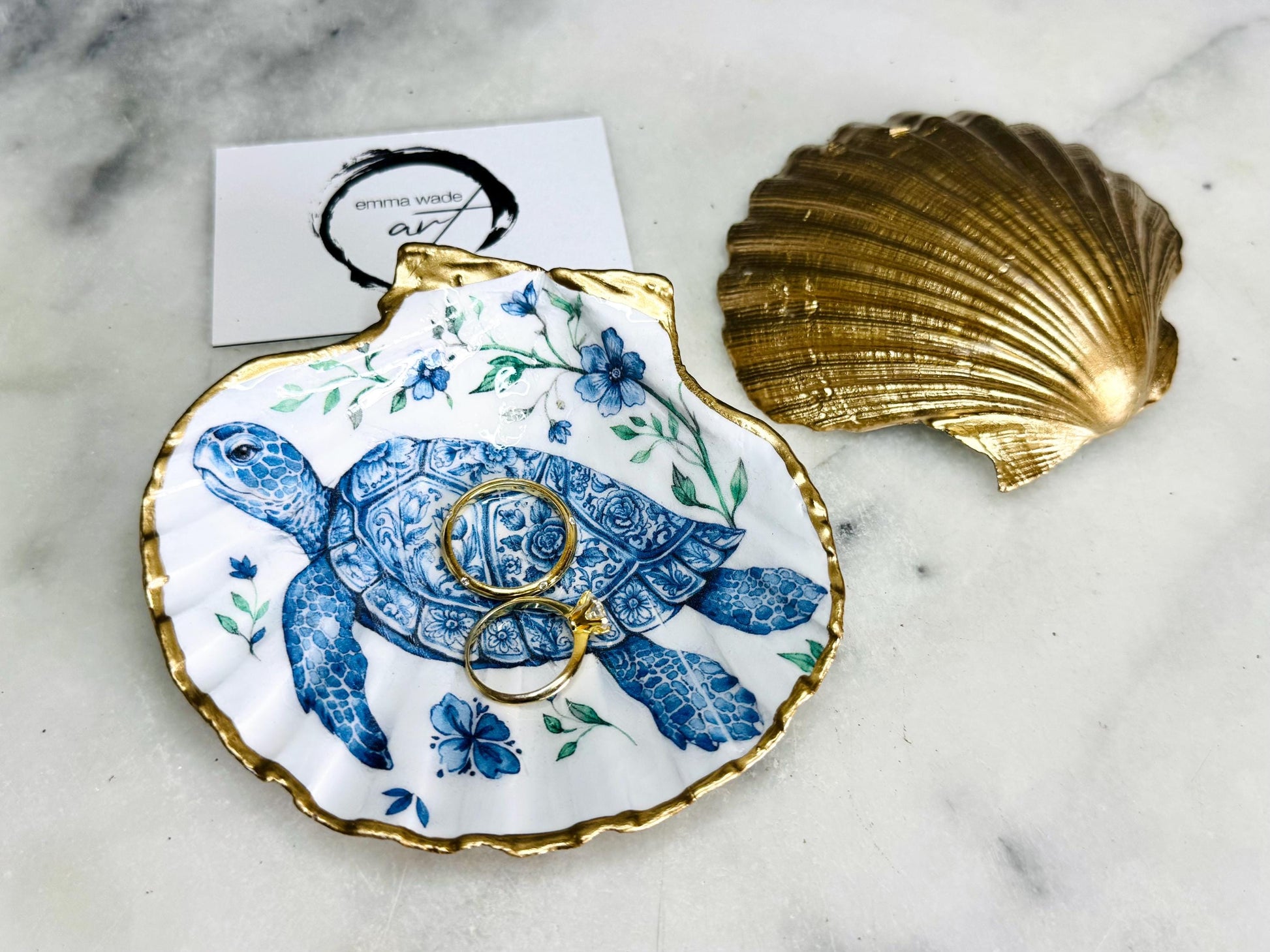 Handcrafted scallop shell trinket dish featuring an original illustrated Delft Blue Sea Turtle design by Emma Wade ART. Natural shell with hand-gilded gold or silver leaf rim and glossy resin finish. Coastal jewellery tray for rings, earrings and keepsakes. Beach house decor, sea life gift and ocean-inspired home accessory available in standard and large sizes.