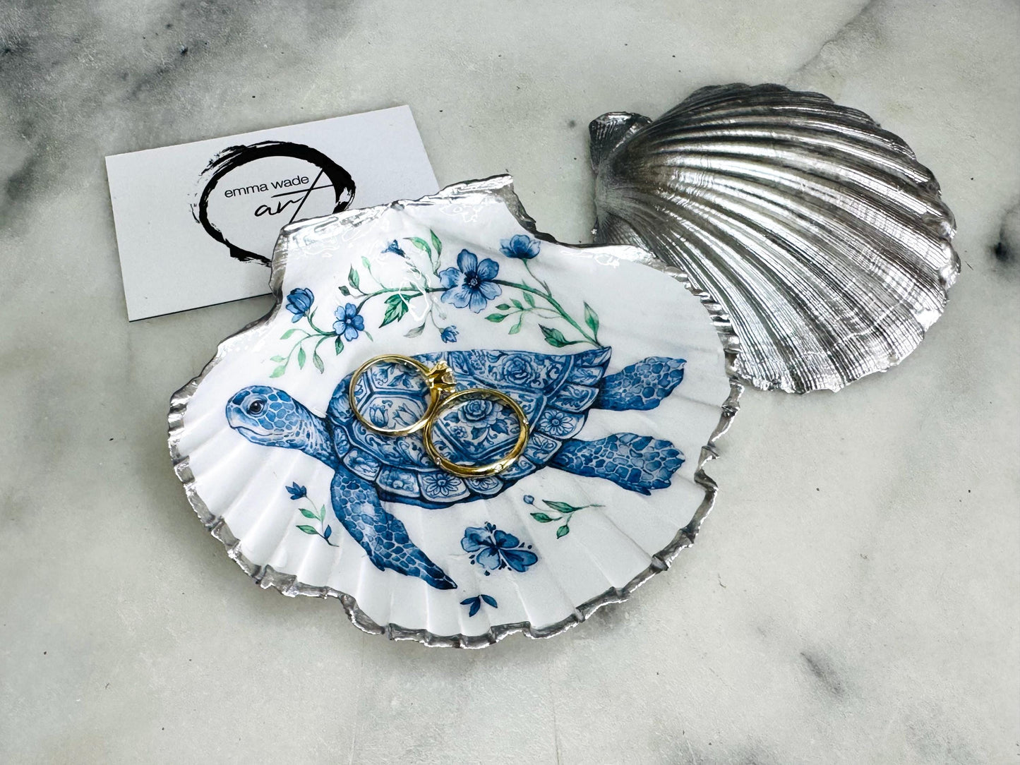 Handcrafted scallop shell trinket dish featuring an original illustrated Delft Blue Sea Turtle design by Emma Wade ART. Natural shell with hand-gilded gold or silver leaf rim and glossy resin finish. Coastal jewellery tray for rings, earrings and keepsakes. Beach house decor, sea life gift and ocean-inspired home accessory available in standard and large sizes.