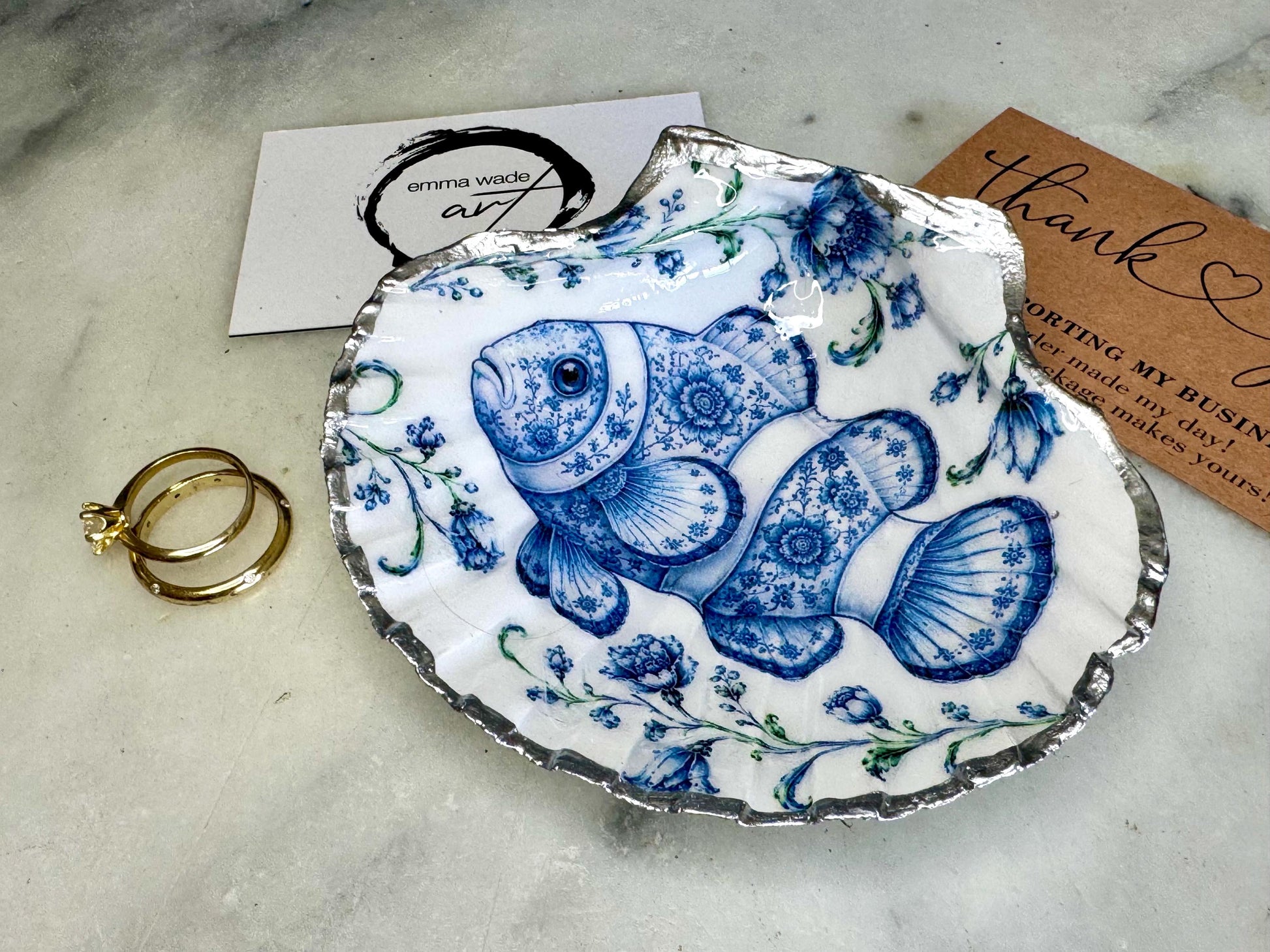 Clownfish scallop shell trinket dish featuring an original illustrated Delft Blue design by Emma Wade ART. Natural scallop shell finished with gold leaf or silver leaf edging and a crystal-clear glossy resin coating. Coastal jewellery tray, ring holder and ocean-inspired gift for fish and sea lovers, beach house décor, weddings, bridesmaids and nautical homes.