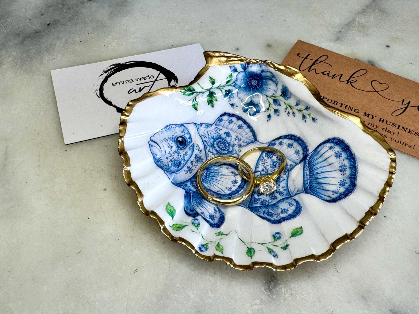 Clownfish scallop shell trinket dish featuring an original illustrated Delft Blue design by Emma Wade ART. Natural scallop shell finished with gold leaf or silver leaf edging and a crystal-clear glossy resin coating. Coastal jewellery tray, Floral ring holder and ocean-inspired gift for fish and sea lovers, beach house décor, weddings, bridesmaids and nautical homes.