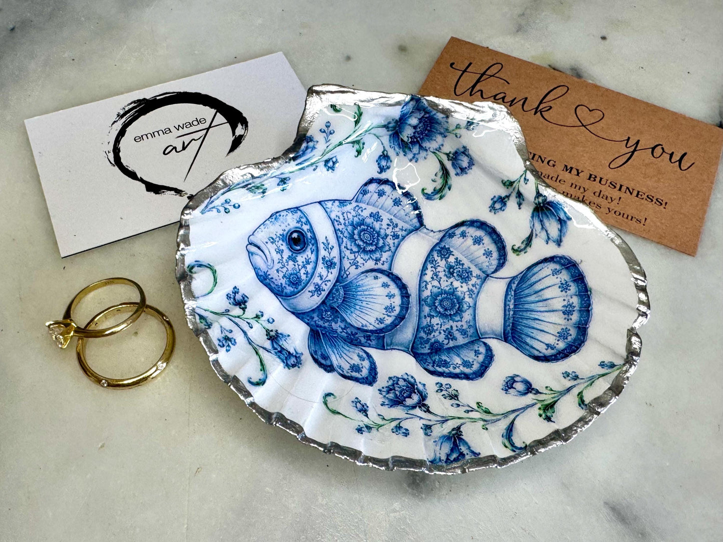 Clownfish scallop shell trinket dish featuring an original illustrated Delft Blue design by Emma Wade ART. Natural scallop shell finished with gold leaf or silver leaf edging and a crystal-clear glossy resin coating. Coastal jewellery tray, Floral ring holder and ocean-inspired gift for fish and sea lovers, beach house décor, weddings, bridesmaids and nautical homes.