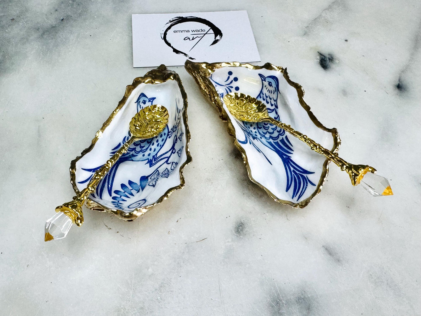 Handmade blue bird oyster shell salt and pepper pinch pot set crafted from natural Dorset oyster shells with glossy pearlescent interiors, blue porcelain style bird artwork and shimmering liquid gold leaf edging. Luxury coastal cruet dishes sealed with gloss resin and styled with optional gold spoons. Elegant seaside dining decor for beach house kitchens, dinner parties, weddings, anniversaries and bird lovers. Artisan shell tableware handmade in the UK with coastal blue and white styling.
