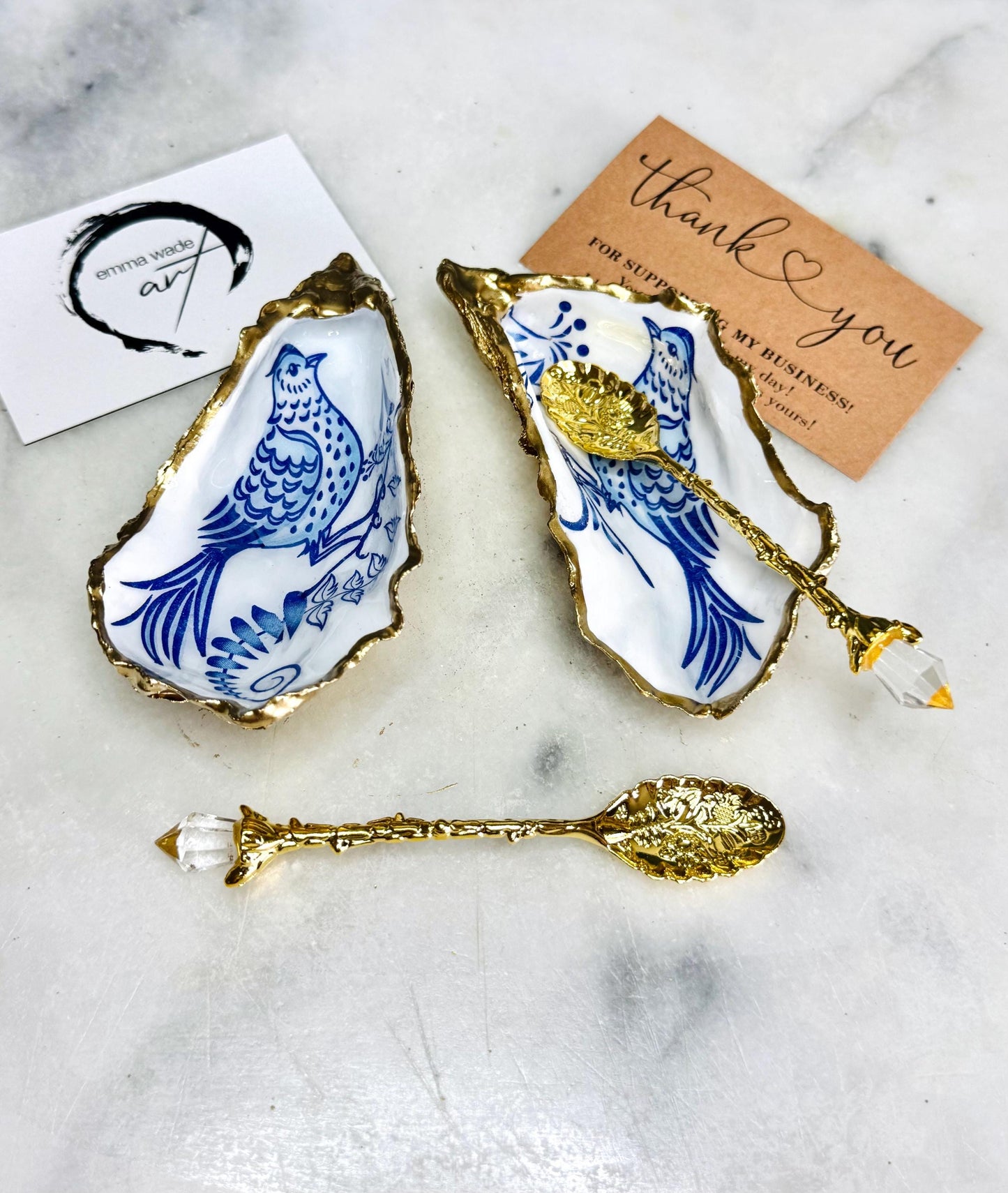 Handmade blue bird oyster shell salt and pepper pinch pot set crafted from natural Dorset oyster shells with glossy pearlescent interiors, blue porcelain style bird artwork and shimmering liquid gold leaf edging. Luxury coastal cruet dishes sealed with gloss resin and styled with optional gold spoons. Elegant seaside dining decor for beach house kitchens, dinner parties, weddings, anniversaries and bird lovers. Artisan shell tableware handmade in the UK with coastal blue and white styling.