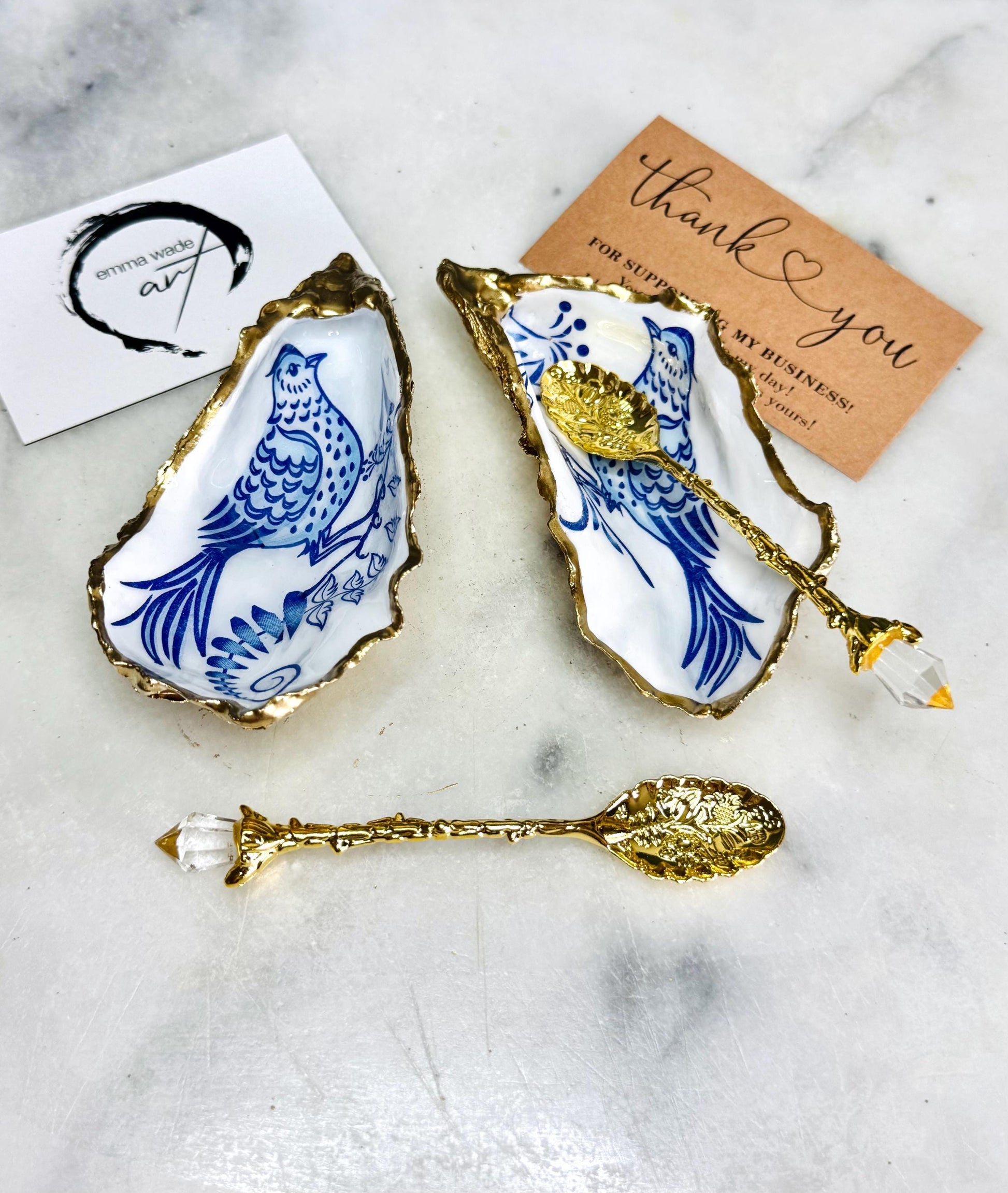Handmade blue bird oyster shell salt and pepper pinch pot set crafted from natural Dorset oyster shells with glossy pearlescent interiors, blue porcelain style bird artwork and shimmering liquid gold leaf edging. Luxury coastal cruet dishes sealed with gloss resin and styled with optional gold spoons. Elegant seaside dining decor for beach house kitchens, dinner parties, weddings, anniversaries and bird lovers. Artisan shell tableware handmade in the UK with coastal blue and white styling.