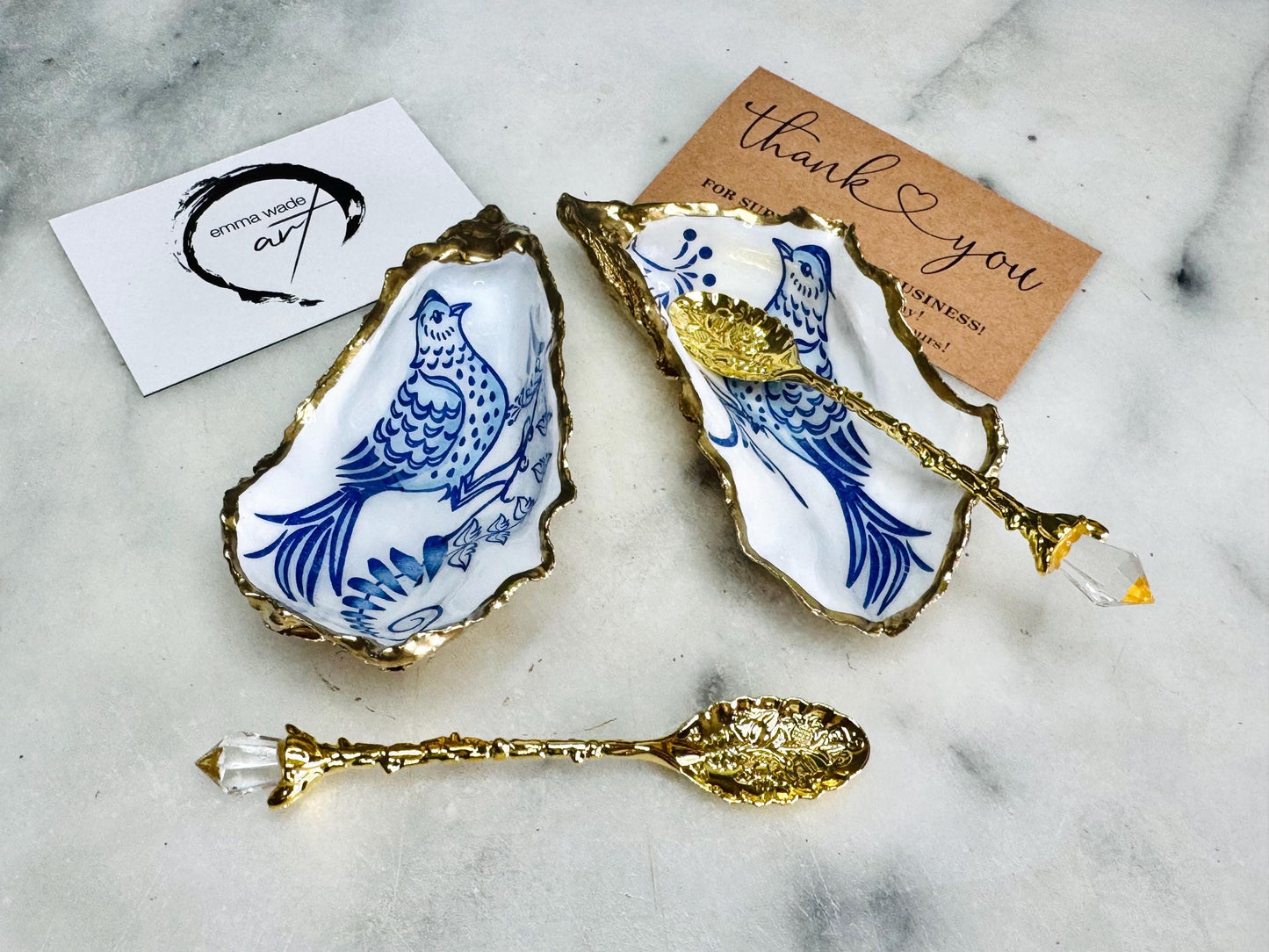 Handmade blue bird oyster shell salt and pepper pinch pot set crafted from natural Dorset oyster shells with glossy pearlescent interiors, blue porcelain style bird artwork and shimmering liquid gold leaf edging. Luxury coastal cruet dishes sealed with gloss resin and styled with optional gold spoons. Elegant seaside dining decor for beach house kitchens, dinner parties, weddings, anniversaries and bird lovers. Artisan shell tableware handmade in the UK with coastal blue and white styling.