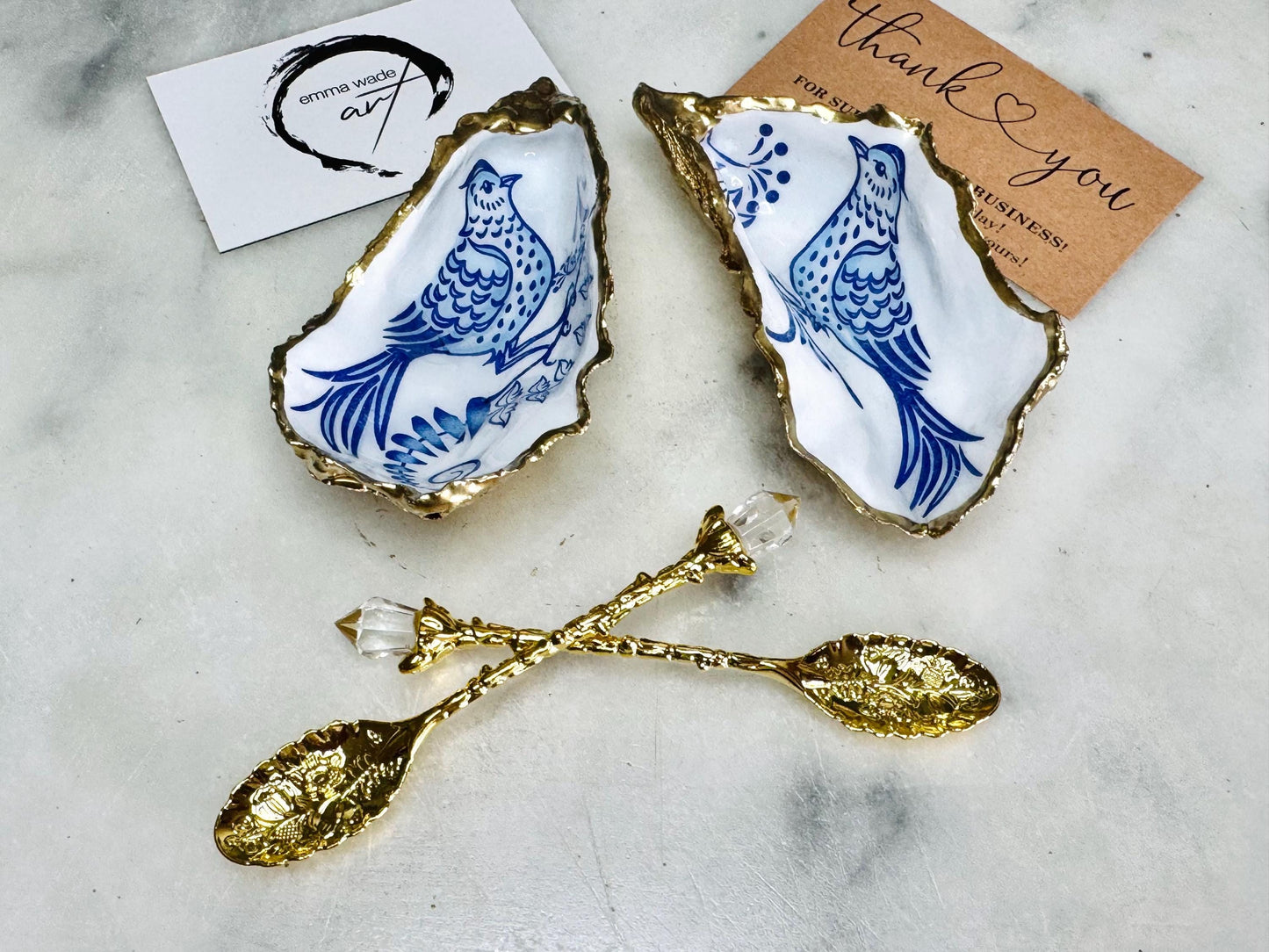 Handmade blue bird oyster shell salt and pepper pinch pot set crafted from natural Dorset oyster shells with glossy pearlescent interiors, blue porcelain style bird artwork and shimmering liquid gold leaf edging. Luxury coastal cruet dishes sealed with gloss resin and styled with optional gold spoons. Elegant seaside dining decor for beach house kitchens, dinner parties, weddings, anniversaries and bird lovers. Artisan shell tableware handmade in the UK with coastal blue and white styling.