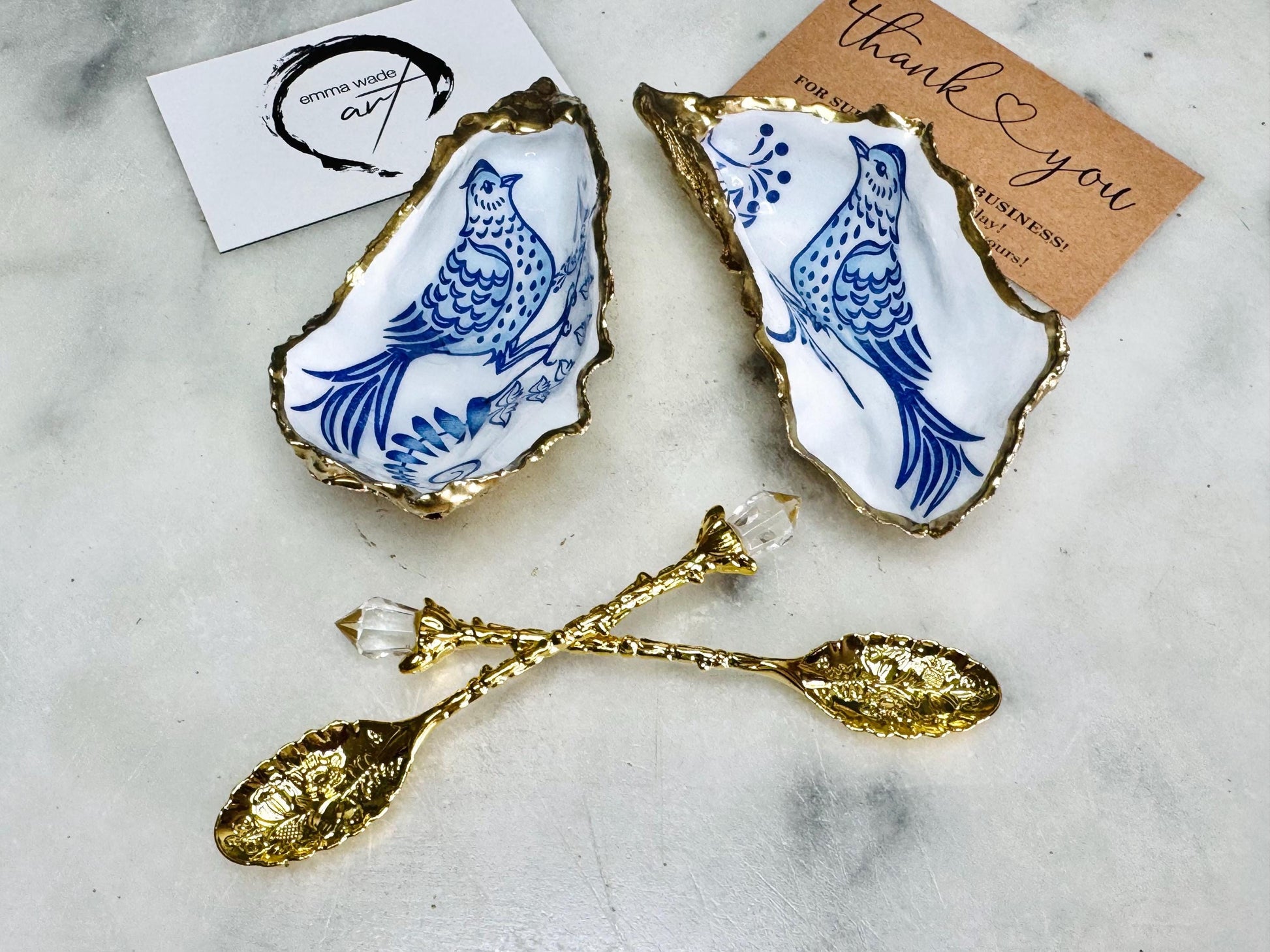 Handmade blue bird oyster shell salt and pepper pinch pot set crafted from natural Dorset oyster shells with glossy pearlescent interiors, blue porcelain style bird artwork and shimmering liquid gold leaf edging. Luxury coastal cruet dishes sealed with gloss resin and styled with optional gold spoons. Elegant seaside dining decor for beach house kitchens, dinner parties, weddings, anniversaries and bird lovers. Artisan shell tableware handmade in the UK with coastal blue and white styling.