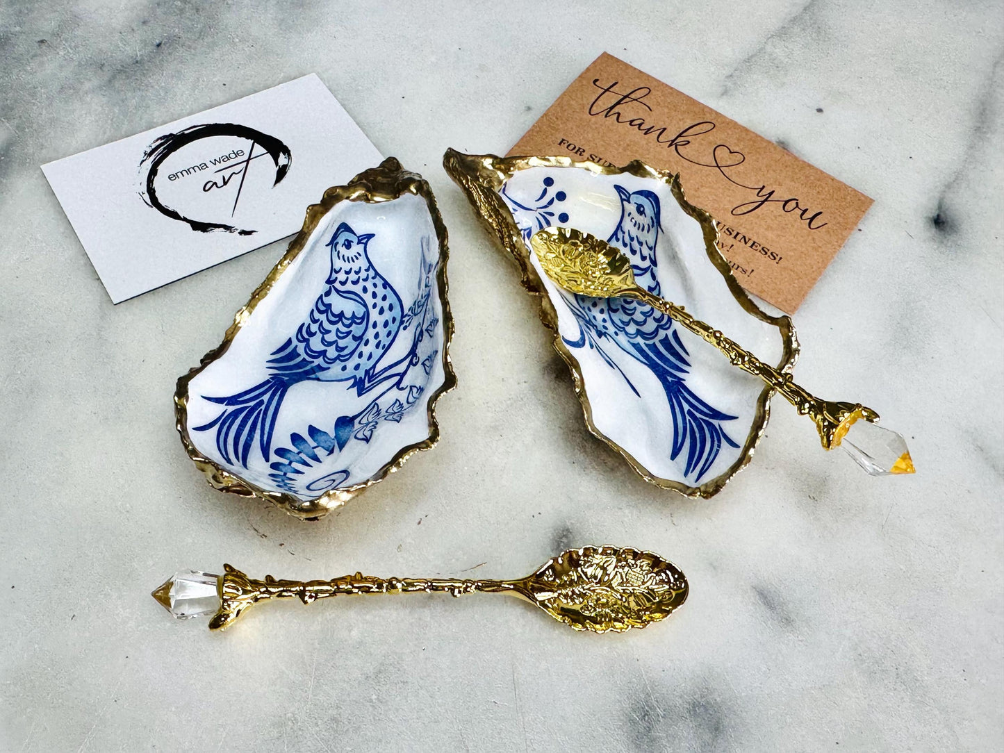 Handmade blue bird oyster shell salt and pepper pinch pot set crafted from natural Dorset oyster shells with glossy pearlescent interiors, blue porcelain style bird artwork and shimmering liquid gold leaf edging. Luxury coastal cruet dishes sealed with gloss resin and styled with optional gold spoons. Elegant seaside dining decor for beach house kitchens, dinner parties, weddings, anniversaries and bird lovers. Artisan shell tableware handmade in the UK with coastal blue and white styling.