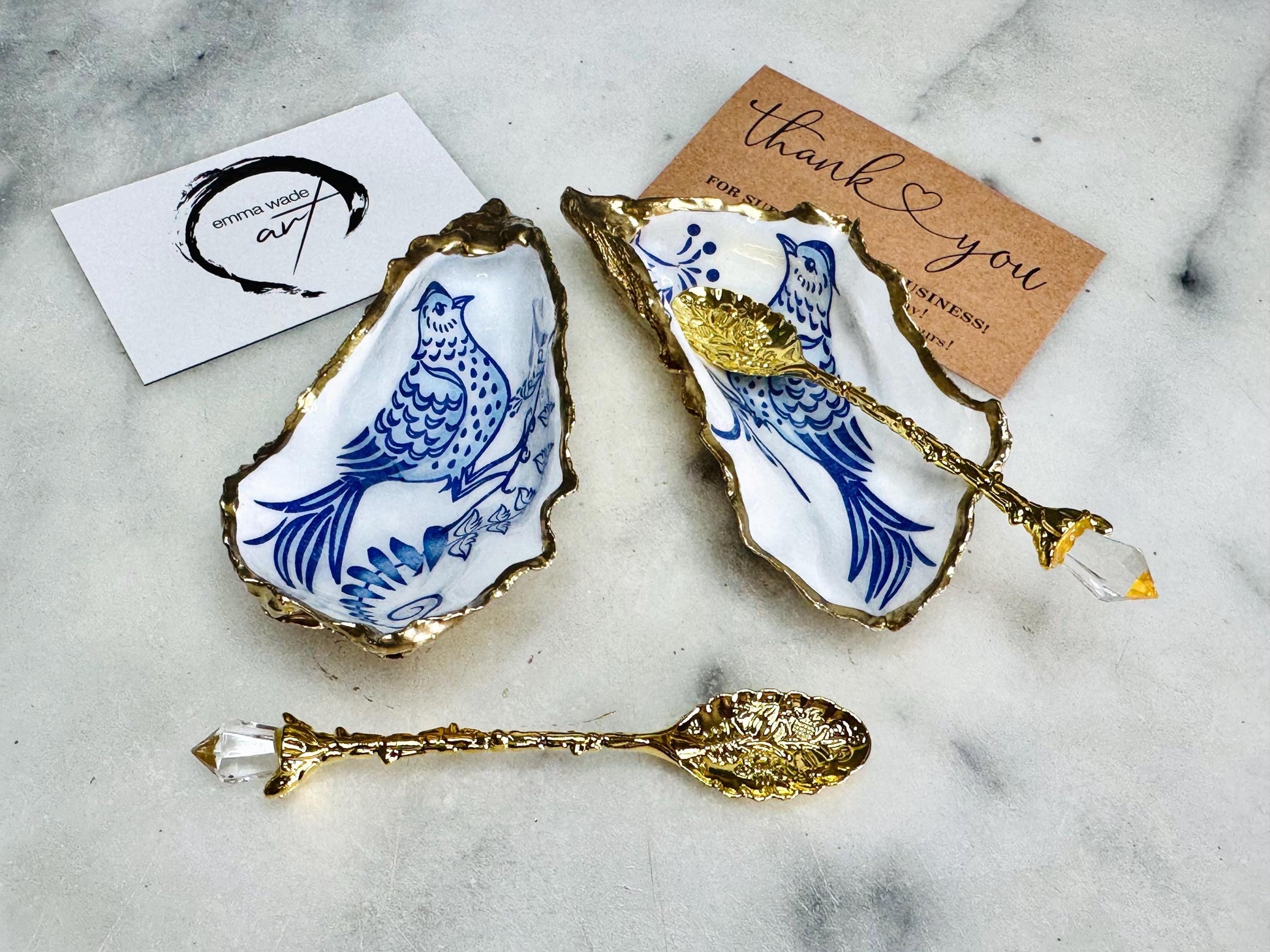 Handmade blue bird oyster shell salt and pepper pinch pot set crafted from natural Dorset oyster shells with glossy pearlescent interiors, blue porcelain style bird artwork and shimmering liquid gold leaf edging. Luxury coastal cruet dishes sealed with gloss resin and styled with optional gold spoons. Elegant seaside dining decor for beach house kitchens, dinner parties, weddings, anniversaries and bird lovers. Artisan shell tableware handmade in the UK with coastal blue and white styling.