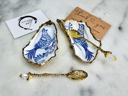 Handmade blue bird oyster shell salt and pepper pinch pot set crafted from natural Dorset oyster shells with glossy pearlescent interiors, blue porcelain style bird artwork and shimmering liquid gold leaf edging. Luxury coastal cruet dishes sealed with gloss resin and styled with optional gold spoons. Elegant seaside dining decor for beach house kitchens, dinner parties, weddings, anniversaries and bird lovers. Artisan shell tableware handmade in the UK with coastal blue and white styling.