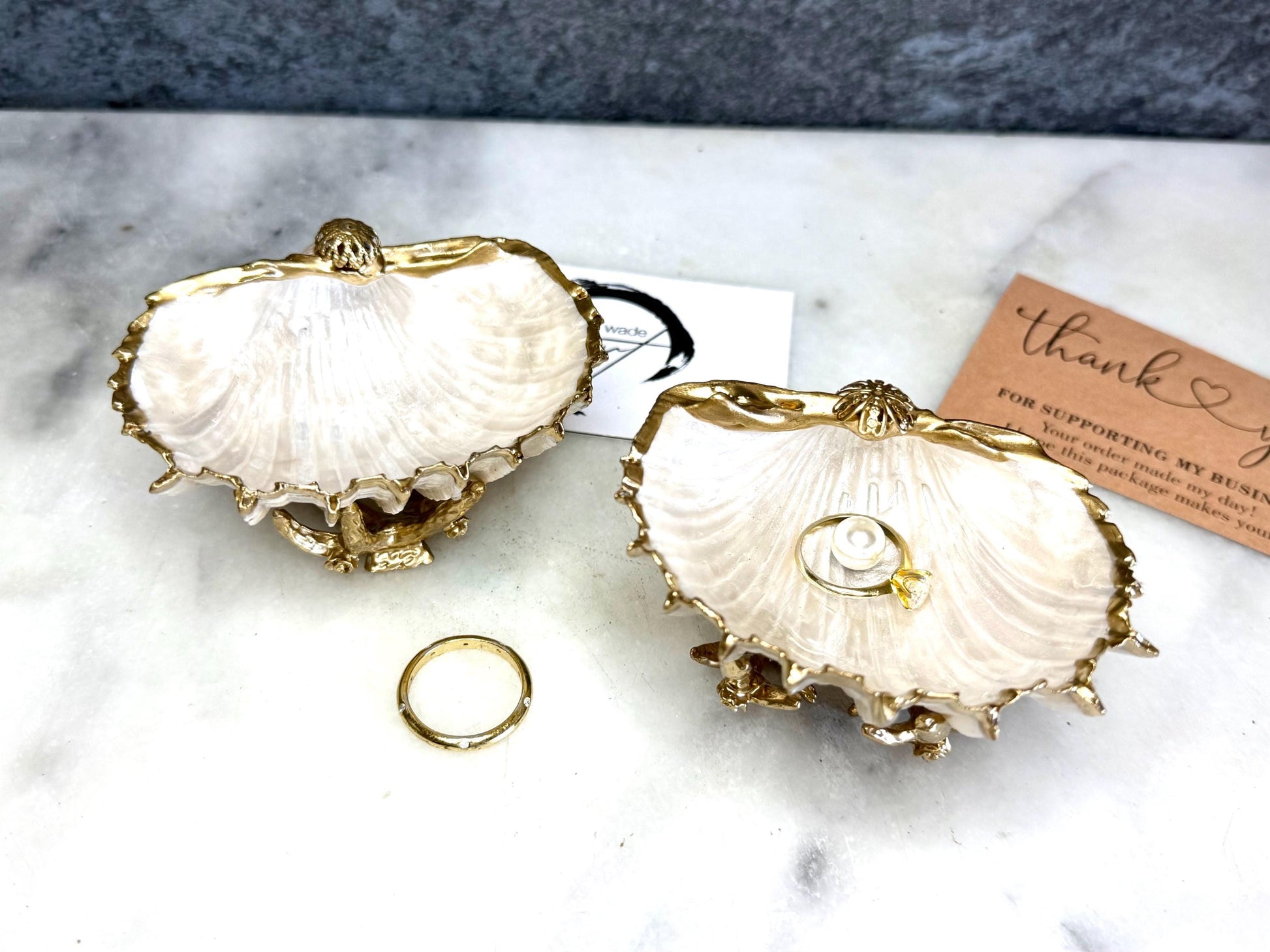 Luxury natural Spiny Cockle shell proposal and wedding ring dish on an ornate gold metal stand, available with or without an inset Swarovski pearl ring holder. Pearlescent shell approximately 8–9cm wide with a hand-gilded liquid gold leaf rim, delicate gold embellishments and crystal-clear resin finish. Handmade luxury jewellery holder, engagement ring dish, bridal keepsake and coastal decor by Emma Wade ART.