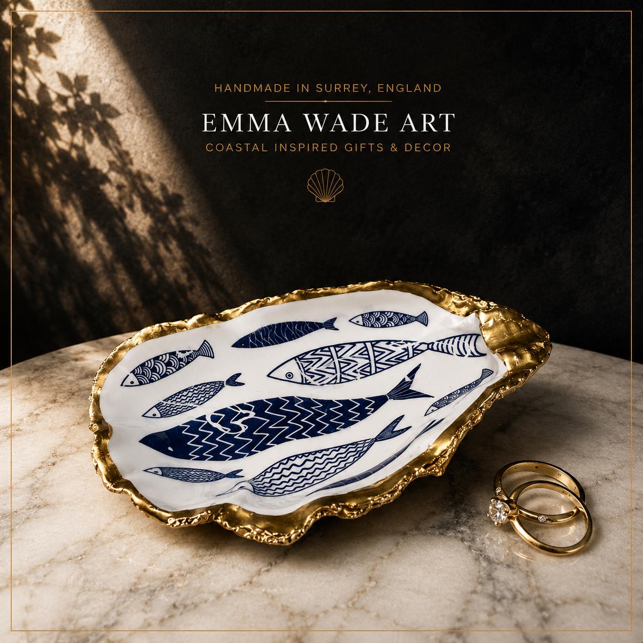 Luxury handmade oyster shell ring dish featuring a blue and white fish design, finished with a hand-gilded liquid gold leaf rim and sealed with crystal-clear resin. Styled on pale marble against a dramatic charcoal background with warm botanical shadows. Coastal jewellery dish, ring holder and decorative shell art handmade in Surrey, England by Emma Wade ART.