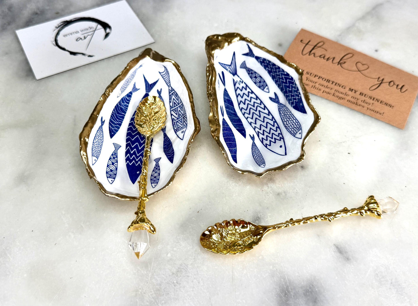 Pair of handmade Dorset oyster shell salt and pepper pinch pots decorated with elegant blue and white fish artwork, finished with hand applied liquid gold leaf edging and sealed in glossy food-safe resin. Coastal shell cruet dishes styled with optional gold crystal spoons on a marble background. Luxury nautical kitchen decor inspired by the sea, perfect for beach house dining, dinner parties, weddings, anniversaries, and coastal home gifts.