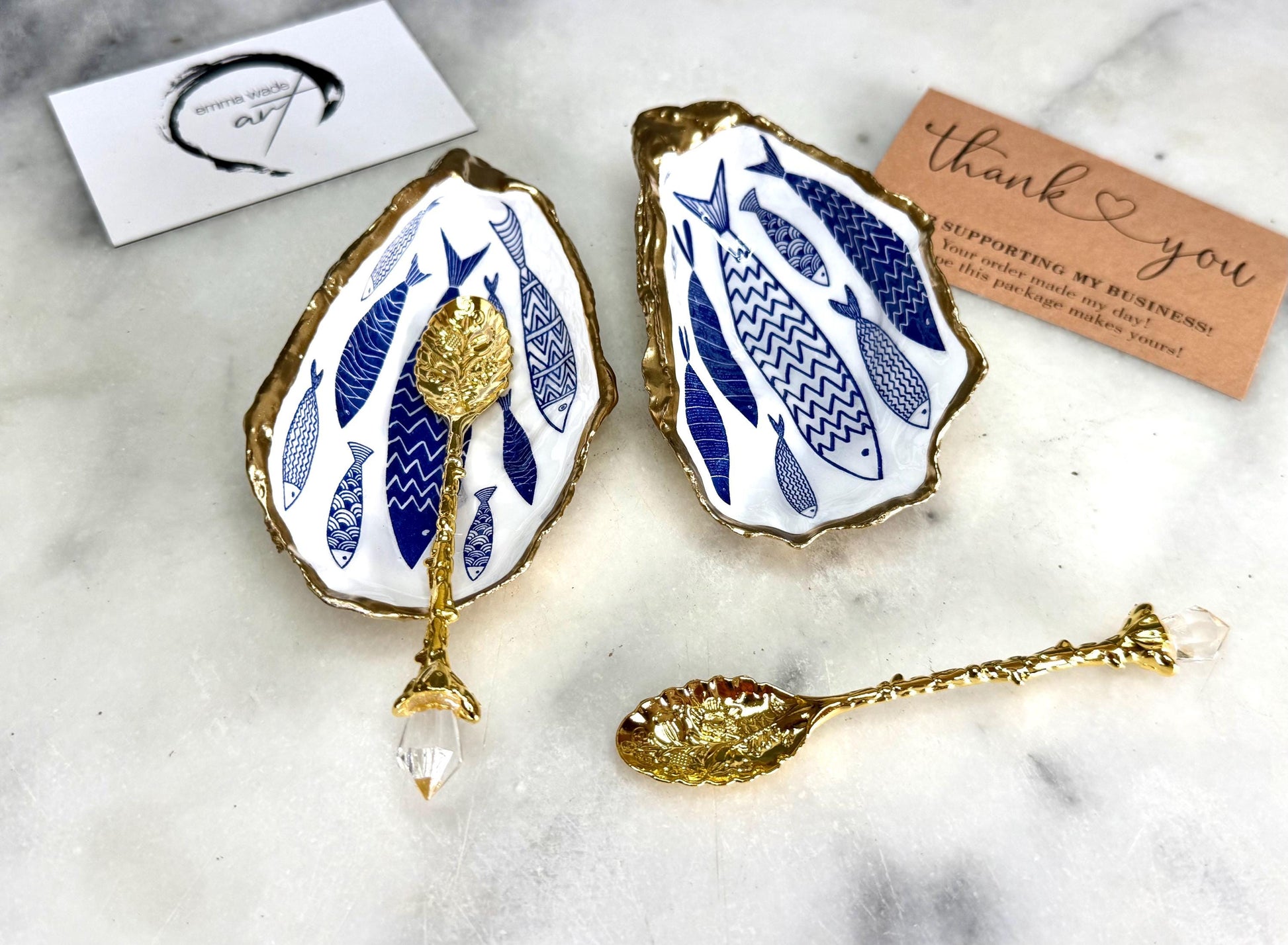 Pair of handmade Dorset oyster shell salt and pepper pinch pots decorated with elegant blue and white fish artwork, finished with hand applied liquid gold leaf edging and sealed in glossy food-safe resin. Coastal shell cruet dishes styled with optional gold crystal spoons on a marble background. Luxury nautical kitchen decor inspired by the sea, perfect for beach house dining, dinner parties, weddings, anniversaries, and coastal home gifts.