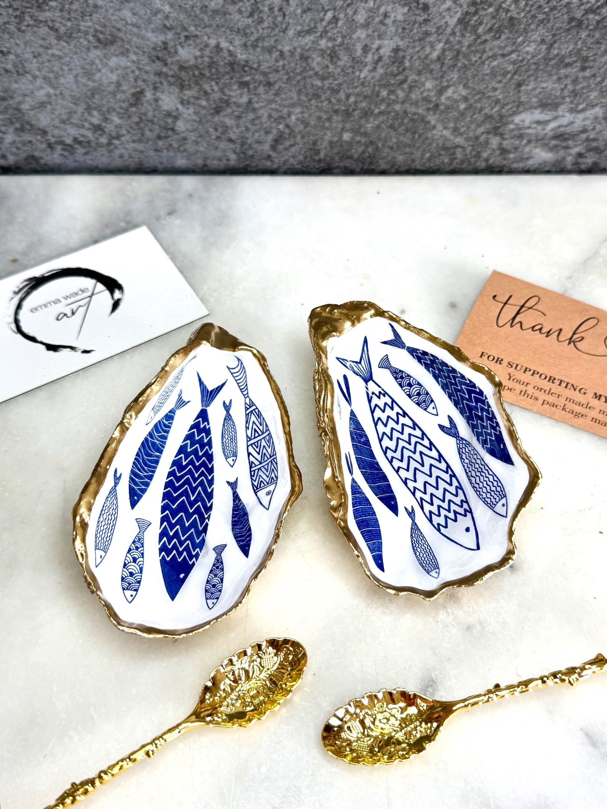 Pair of handmade Dorset oyster shell salt and pepper pinch pots decorated with elegant blue and white fish artwork, finished with hand applied liquid gold leaf edging and sealed in glossy food-safe resin. Coastal shell cruet dishes styled with optional gold crystal spoons on a marble background. Luxury nautical kitchen decor inspired by the sea, perfect for beach house dining, dinner parties, weddings, anniversaries, and coastal home gifts.