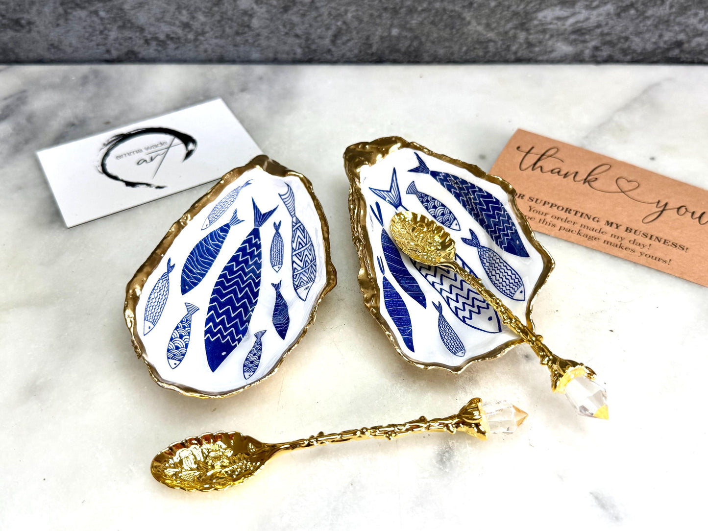 Pair of handmade Dorset oyster shell salt and pepper pinch pots decorated with elegant blue and white fish artwork, finished with hand applied liquid gold leaf edging and sealed in glossy food-safe resin. Coastal shell cruet dishes styled with optional gold crystal spoons on a marble background. Luxury nautical kitchen decor inspired by the sea, perfect for beach house dining, dinner parties, weddings, anniversaries, and coastal home gifts.