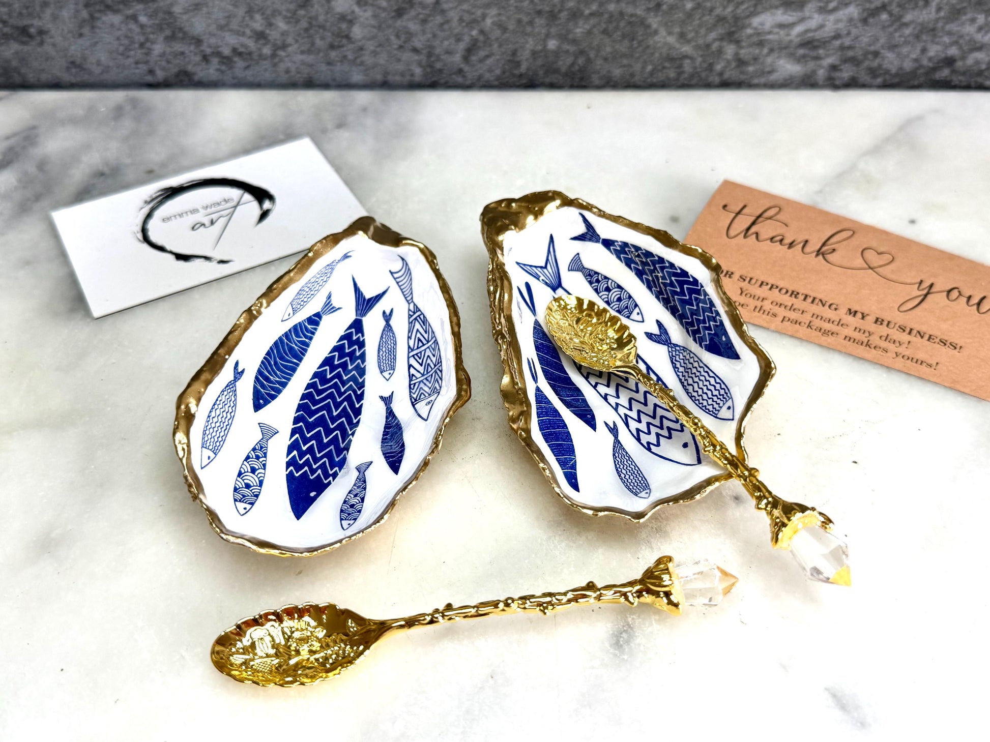 Pair of handmade Dorset oyster shell salt and pepper pinch pots decorated with elegant blue and white fish artwork, finished with hand applied liquid gold leaf edging and sealed in glossy food-safe resin. Coastal shell cruet dishes styled with optional gold crystal spoons on a marble background. Luxury nautical kitchen decor inspired by the sea, perfect for beach house dining, dinner parties, weddings, anniversaries, and coastal home gifts.