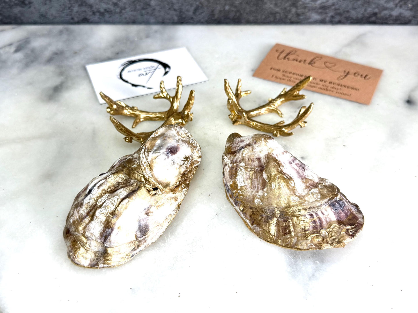 Pair of natural cupped oyster shell salt and pepper dishes displayed on individual driftwood-style gold metal stands. Each oyster shell has a liquid gold leaf rim and glossy resin-sealed pearlescent interior, with a choice of natural gold-highlighted or fully gilded backs. Optional ornate gold spoons with clear crystal ends are shown alongside. Luxury coastal cruet set and wedding or housewarming gift by Emma Wade ART.