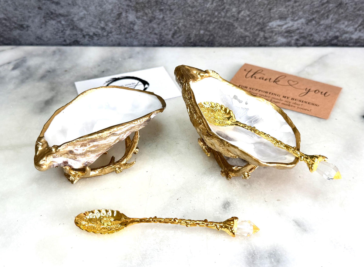 Pair of natural cupped oyster shell salt and pepper dishes displayed on individual driftwood-style gold metal stands. Each oyster shell has a liquid gold leaf rim and glossy resin-sealed pearlescent interior, with a choice of natural gold-highlighted or fully gilded backs. Optional ornate gold spoons with clear crystal ends are shown alongside. Luxury coastal cruet set and wedding or housewarming gift by Emma Wade ART.