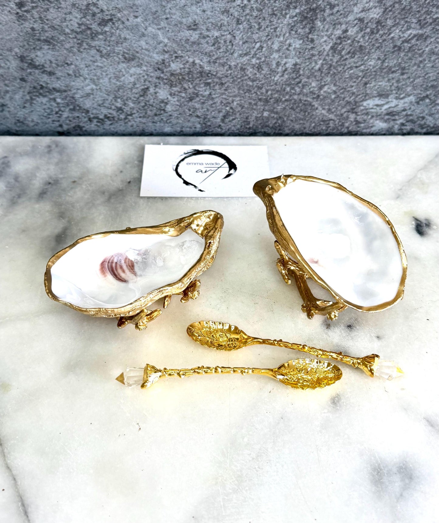 Pair of natural cupped oyster shell salt and pepper dishes displayed on individual driftwood-style gold metal stands. Each oyster shell has a liquid gold leaf rim and glossy resin-sealed pearlescent interior, with a choice of natural gold-highlighted or fully gilded backs. Optional ornate gold spoons with clear crystal ends are shown alongside. Luxury coastal cruet set and wedding or housewarming gift by Emma Wade ART.
