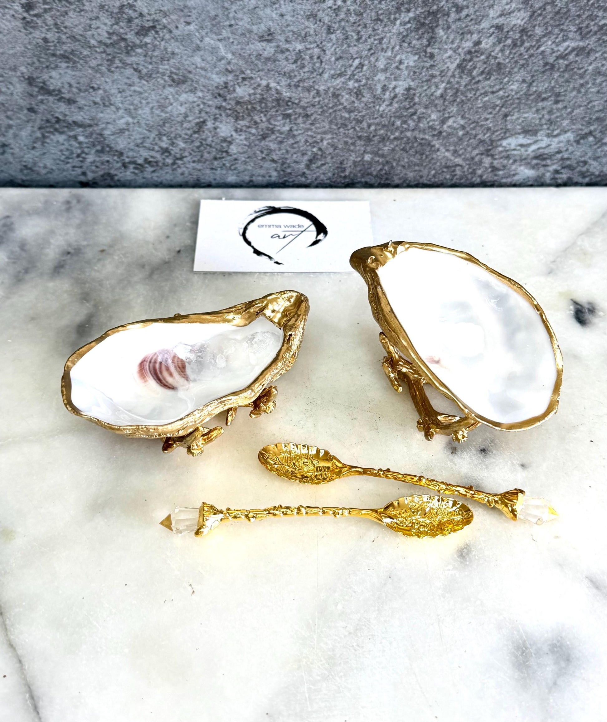 Pair of natural cupped oyster shell salt and pepper dishes displayed on individual driftwood-style gold metal stands. Each oyster shell has a liquid gold leaf rim and glossy resin-sealed pearlescent interior, with a choice of natural gold-highlighted or fully gilded backs. Optional ornate gold spoons with clear crystal ends are shown alongside. Luxury coastal cruet set and wedding or housewarming gift by Emma Wade ART.