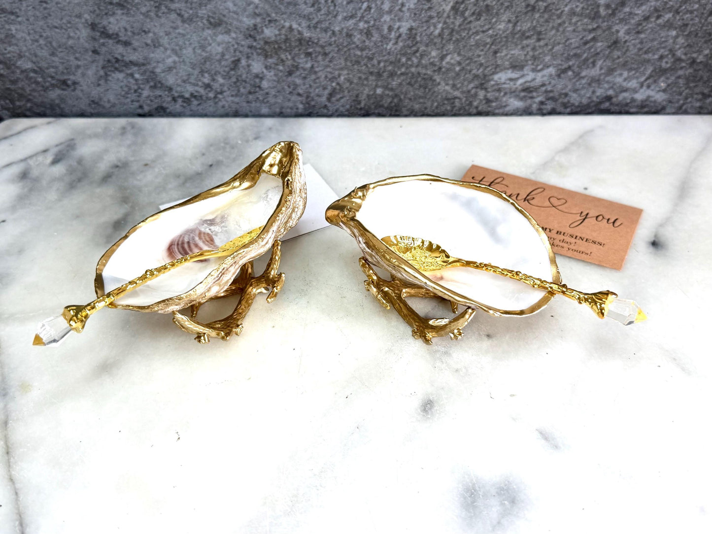 Pair of natural cupped oyster shell salt and pepper dishes displayed on individual driftwood-style gold metal stands. Each oyster shell has a liquid gold leaf rim and glossy resin-sealed pearlescent interior, with a choice of natural gold-highlighted or fully gilded backs. Optional ornate gold spoons with clear crystal ends are shown alongside. Luxury coastal cruet set and wedding or housewarming gift by Emma Wade ART.