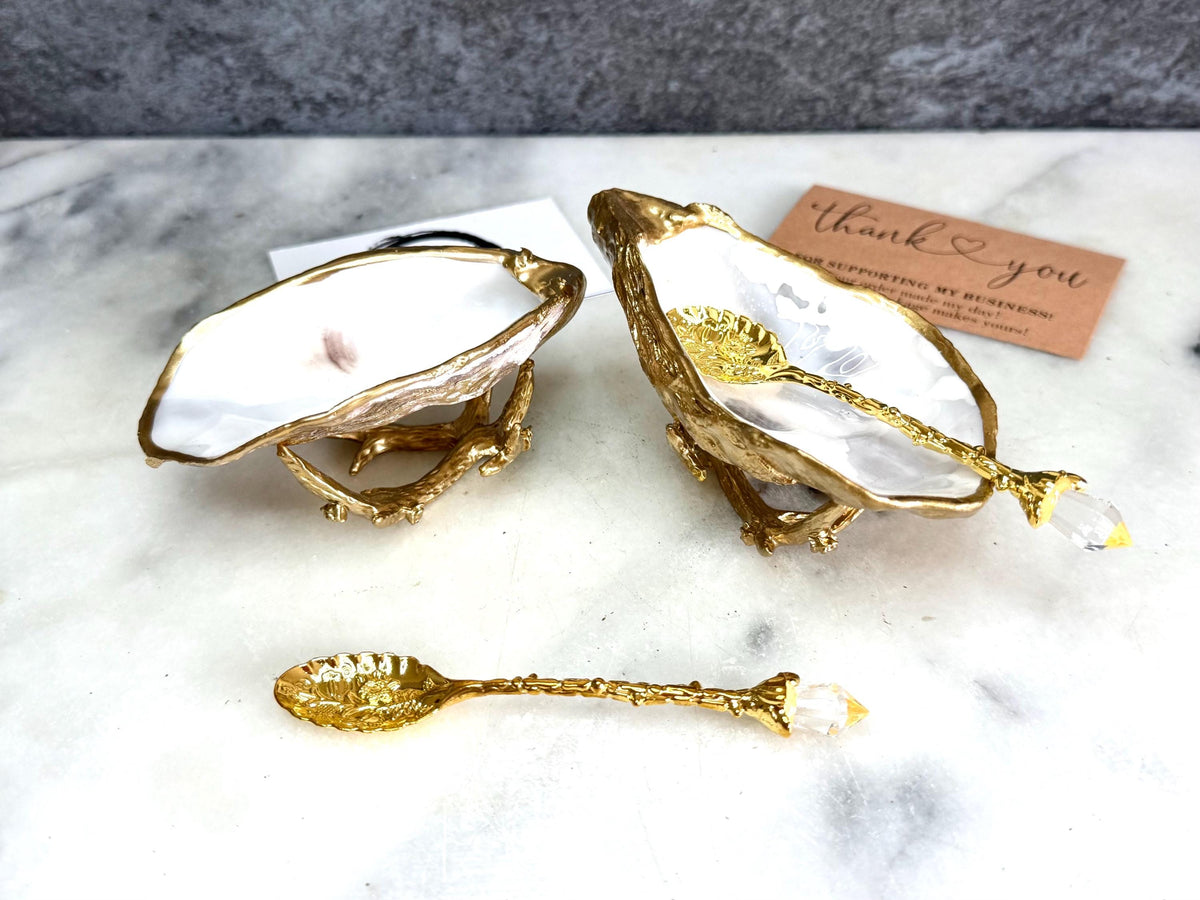 Pair of natural cupped oyster shell salt and pepper dishes displayed on individual driftwood-style gold metal stands. Each oyster shell has a liquid gold leaf rim and glossy resin-sealed pearlescent interior, with a choice of natural gold-highlighted or fully gilded backs. Optional ornate gold spoons with clear crystal ends are shown alongside. Luxury coastal cruet set and wedding or housewarming gift by Emma Wade ART.