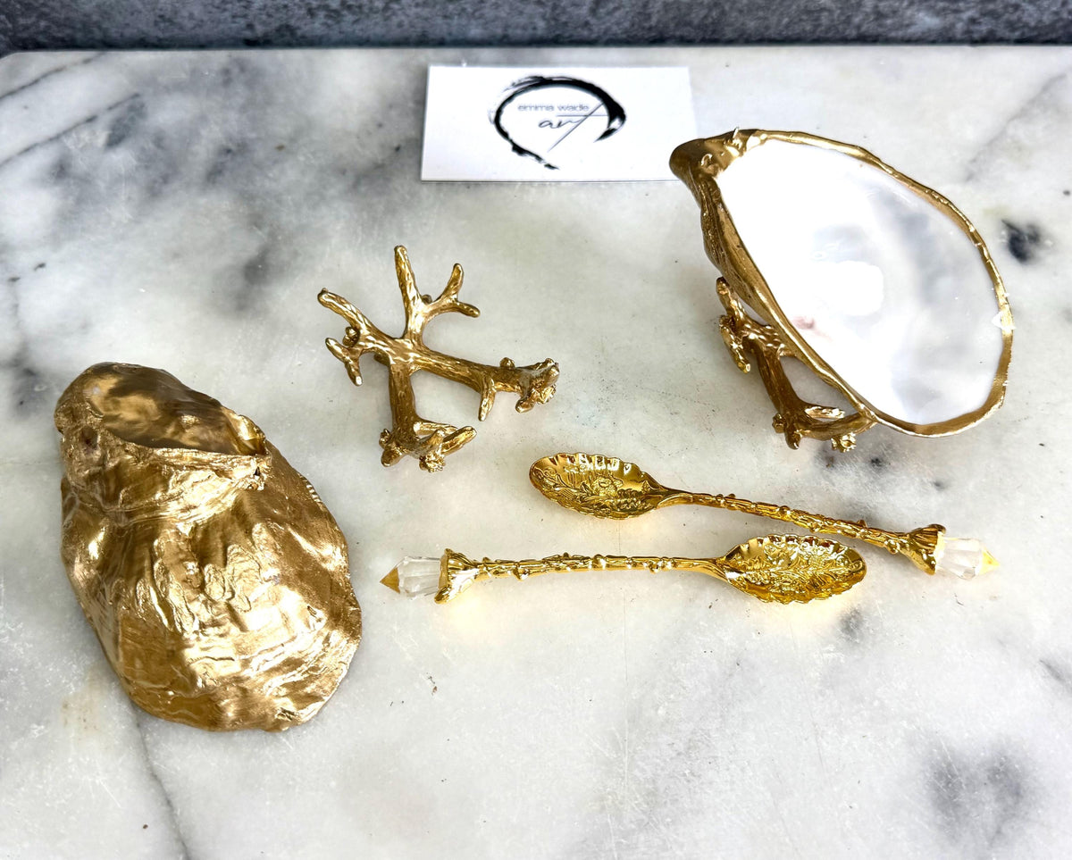 Pair of natural cupped oyster shell salt and pepper dishes displayed on individual driftwood-style gold metal stands. Each oyster shell has a liquid gold leaf rim and glossy resin-sealed pearlescent interior, with a choice of natural gold-highlighted or fully gilded backs. Optional ornate gold spoons with clear crystal ends are shown alongside. Luxury coastal cruet set and wedding or housewarming gift by Emma Wade ART.