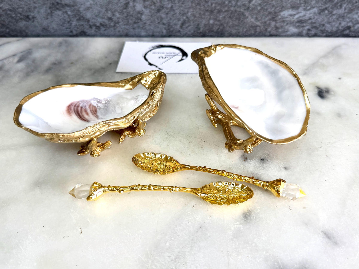 Pair of natural cupped oyster shell salt and pepper dishes displayed on individual driftwood-style gold metal stands. Each oyster shell has a liquid gold leaf rim and glossy resin-sealed pearlescent interior, with a choice of natural gold-highlighted or fully gilded backs. Optional ornate gold spoons with clear crystal ends are shown alongside. Luxury coastal cruet set and wedding or housewarming gift by Emma Wade ART.