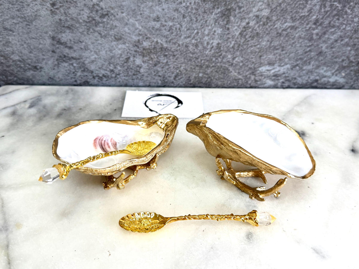 Pair of natural cupped oyster shell salt and pepper dishes displayed on individual driftwood-style gold metal stands. Each oyster shell has a liquid gold leaf rim and glossy resin-sealed pearlescent interior, with a choice of natural gold-highlighted or fully gilded backs. Optional ornate gold spoons with clear crystal ends are shown alongside. Luxury coastal cruet set and wedding or housewarming gift by Emma Wade ART.
