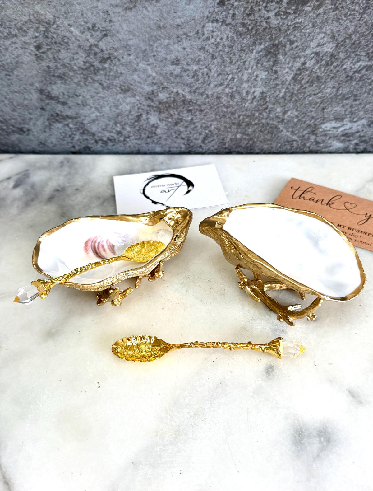 Pair of natural cupped oyster shell salt and pepper dishes displayed on individual driftwood-style gold metal stands. Each oyster shell has a liquid gold leaf rim and glossy resin-sealed pearlescent interior, with a choice of natural gold-highlighted or fully gilded backs. Optional ornate gold spoons with clear crystal ends are shown alongside. Luxury coastal cruet set and wedding or housewarming gift by Emma Wade ART.