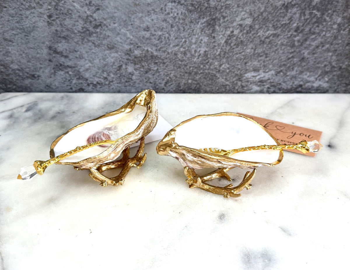 Pair of natural cupped oyster shell salt and pepper dishes displayed on individual driftwood-style gold metal stands. Each oyster shell has a liquid gold leaf rim and glossy resin-sealed pearlescent interior, with a choice of natural gold-highlighted or fully gilded backs. Optional ornate gold spoons with clear crystal ends are shown alongside. Luxury coastal cruet set and wedding or housewarming gift by Emma Wade ART.