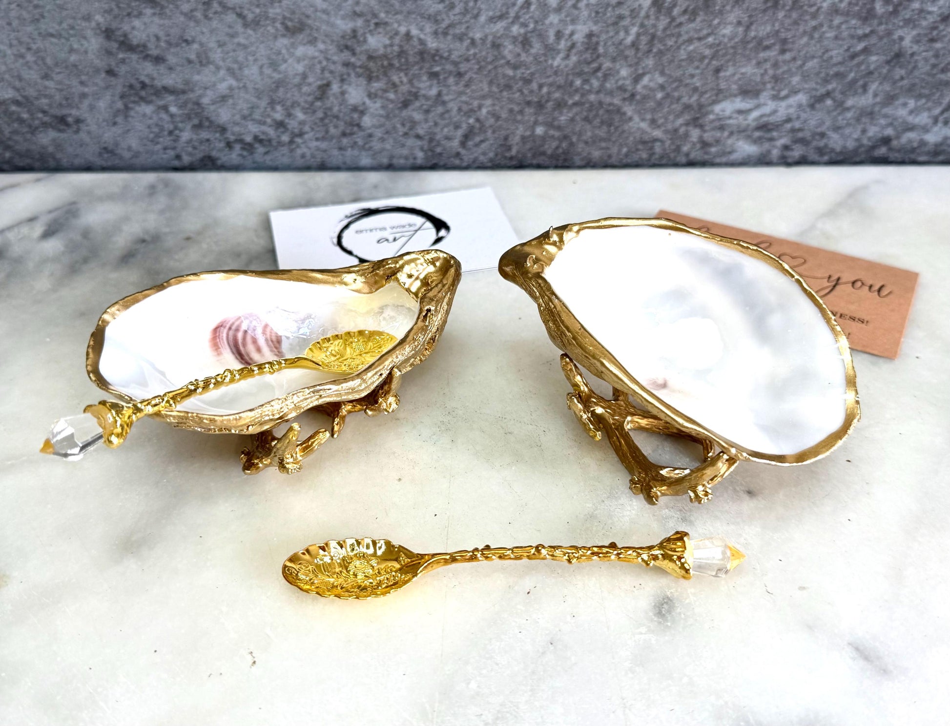 Pair of natural cupped oyster shell salt and pepper dishes displayed on individual driftwood-style gold metal stands. Each oyster shell has a liquid gold leaf rim and glossy resin-sealed pearlescent interior, with a choice of natural gold-highlighted or fully gilded backs. Optional ornate gold spoons with clear crystal ends are shown alongside. Luxury coastal cruet set and wedding or housewarming gift by Emma Wade ART.