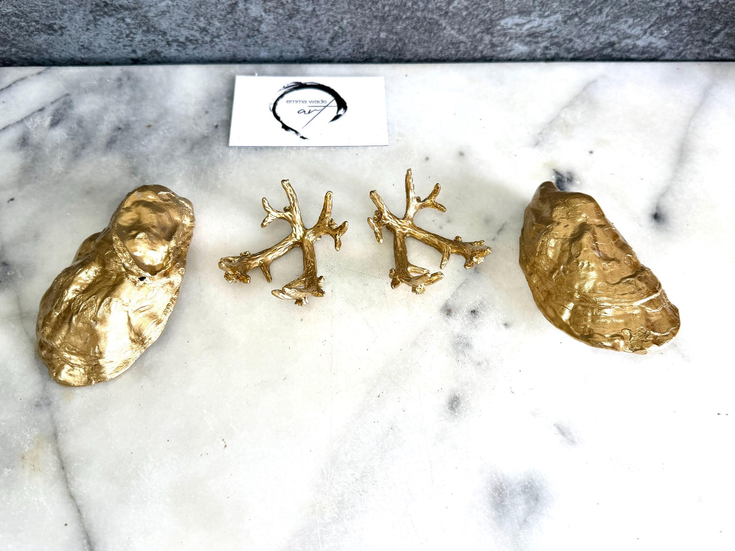 Pair of natural cupped oyster shell salt and pepper dishes displayed on individual driftwood-style gold metal stands. Each oyster shell has a liquid gold leaf rim and glossy resin-sealed pearlescent interior, with a choice of natural gold-highlighted or fully gilded backs. Optional ornate gold spoons with clear crystal ends are shown alongside. Luxury coastal cruet set and wedding or housewarming gift by Emma Wade ART.