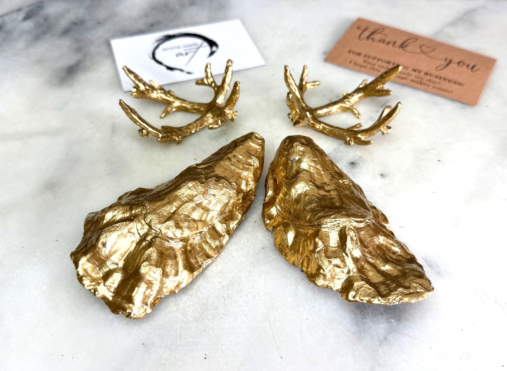 Pair of natural cupped oyster shell salt and pepper dishes displayed on individual driftwood-style gold metal stands. Each oyster shell has a liquid gold leaf rim and glossy resin-sealed pearlescent interior, with a choice of natural gold-highlighted or fully gilded backs. Optional ornate gold spoons with clear crystal ends are shown alongside. Luxury coastal cruet set and wedding or housewarming gift by Emma Wade ART.
