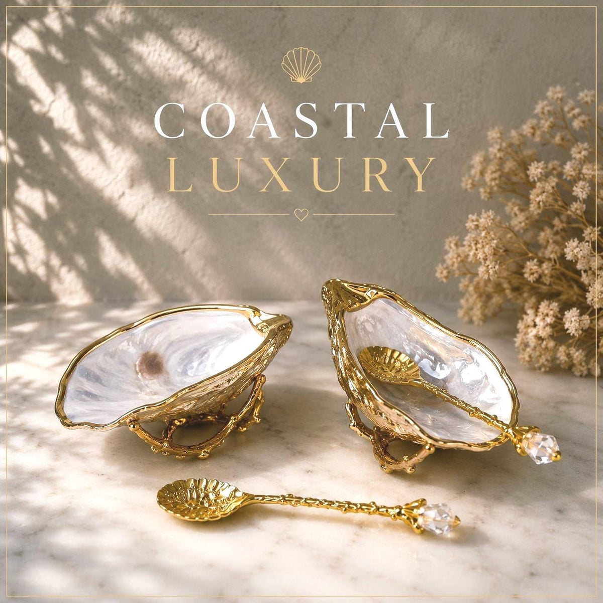 Pair of natural cupped oyster shell salt and pepper dishes displayed on individual driftwood-style gold metal stands. Each oyster shell has a liquid gold leaf rim and glossy resin-sealed pearlescent interior, with a choice of natural gold-highlighted or fully gilded backs. Optional ornate gold spoons with clear crystal ends are shown alongside. Luxury coastal cruet set and wedding or housewarming gift by Emma Wade ART.