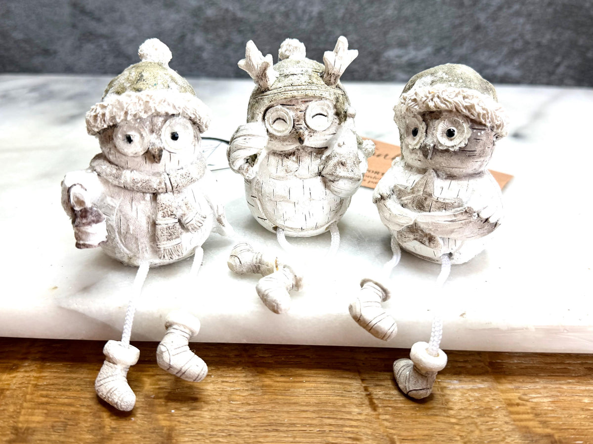 Set of three woodland Christmas owl shelf sitter decorations in soft white, grey and natural tones, featuring winter hats, scarves and festive details. Moulded resin owl ornaments with subtle glitter accents and dangling cord legs with little boots, designed to sit on a shelf, mantelpiece or windowsill. Festive woodland Christmas décor and owl lover gift.