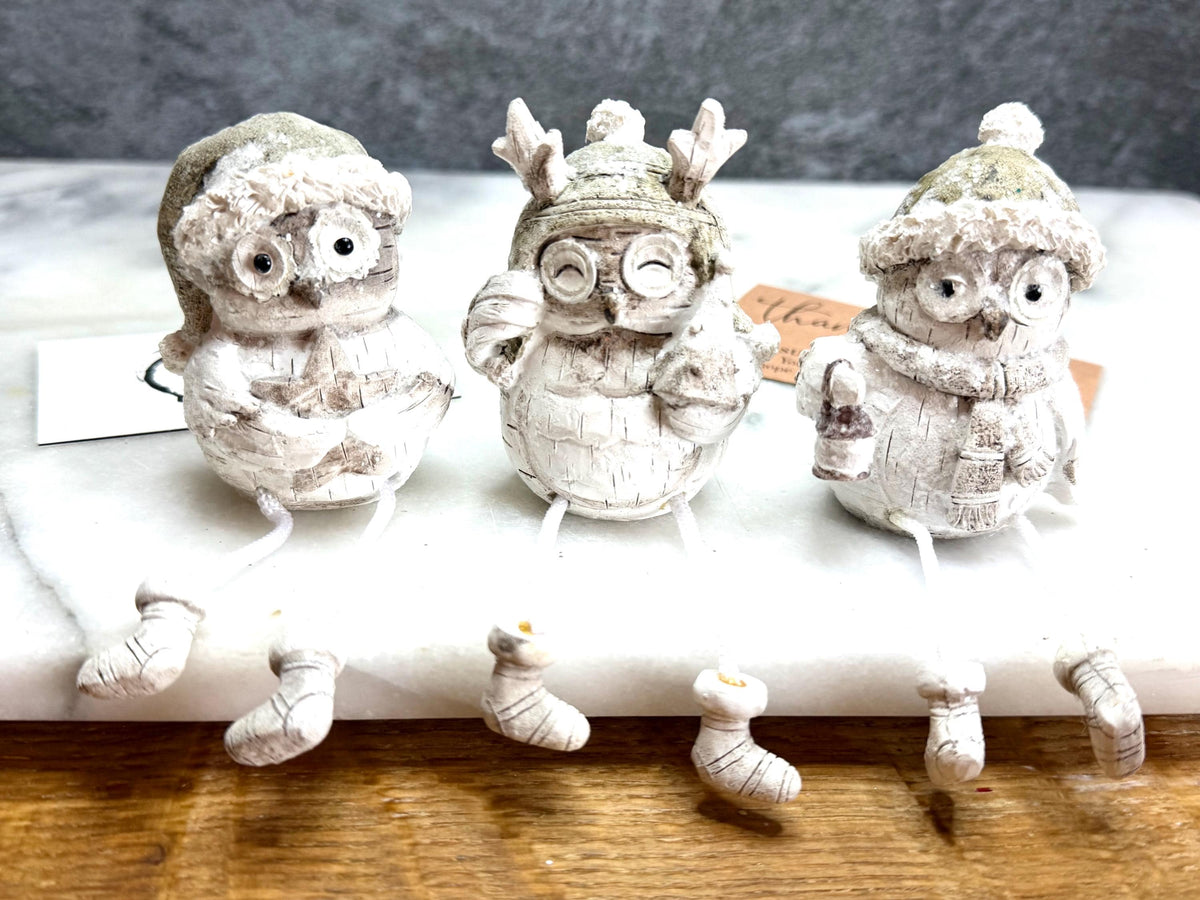 Set of three woodland Christmas owl shelf sitter decorations in soft white, grey and natural tones, featuring winter hats, scarves and festive details. Moulded resin owl ornaments with subtle glitter accents and dangling cord legs with little boots, designed to sit on a shelf, mantelpiece or windowsill. Festive woodland Christmas décor and owl lover gift.
